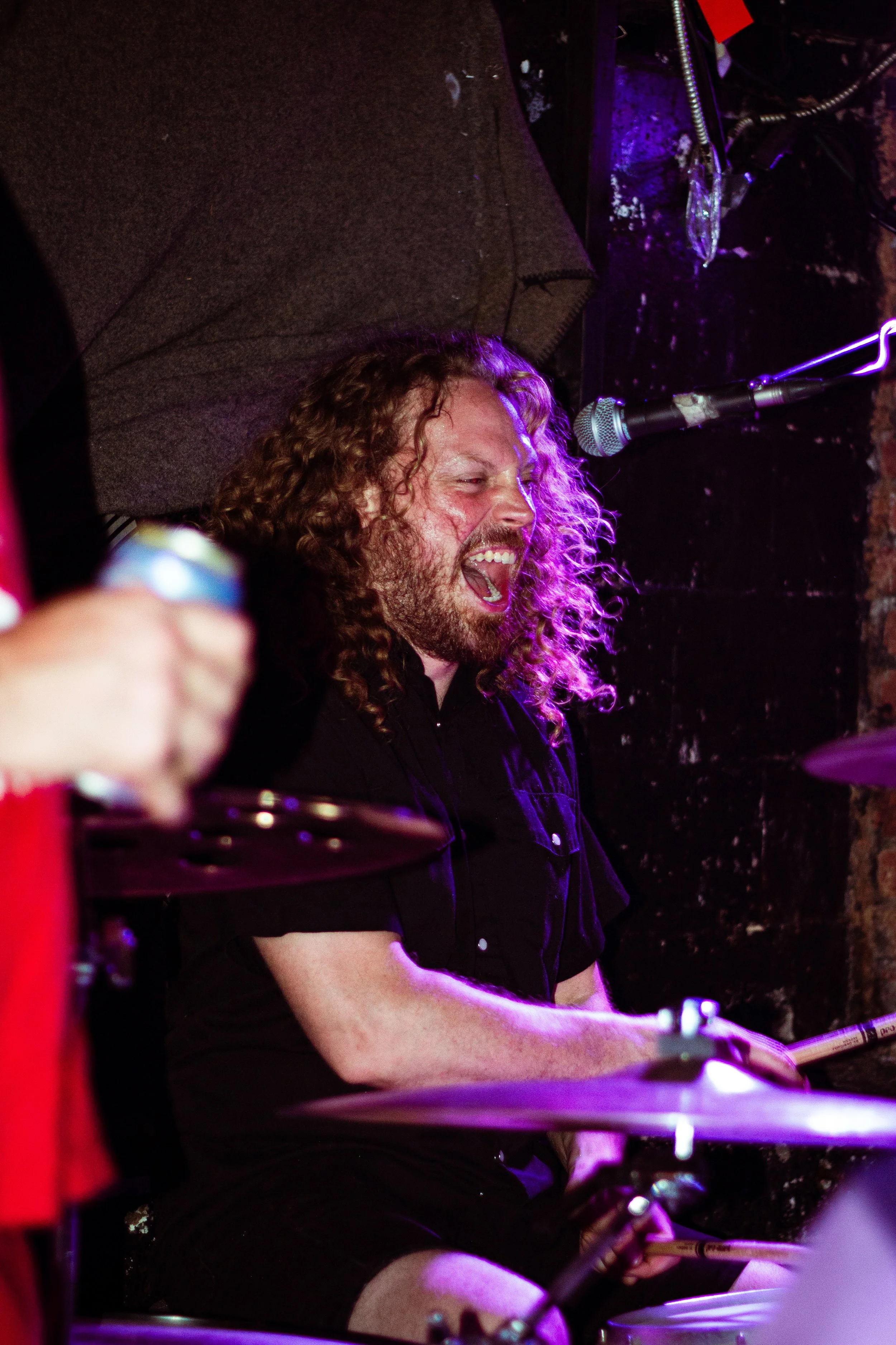 A man with long, curly hair and a beard passionately singing or shouting while playing drums on a dark stage with purple lighting.