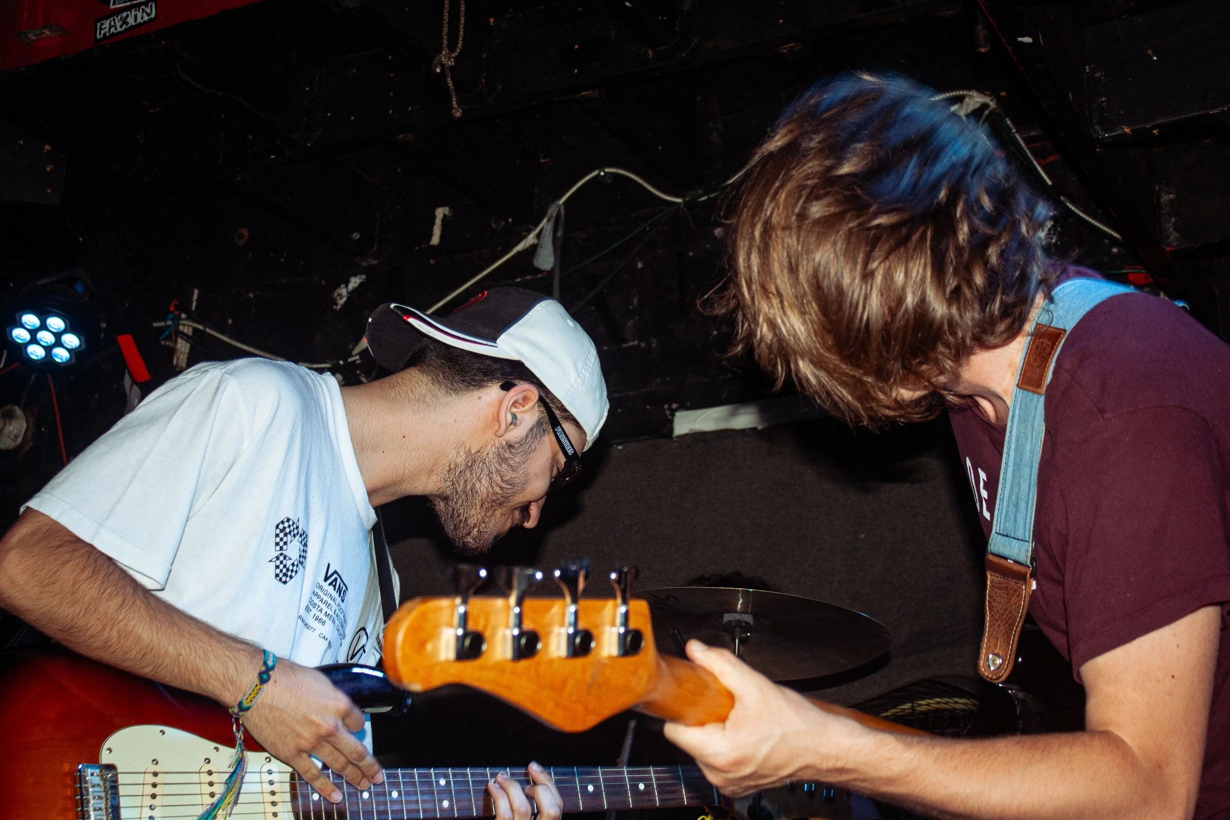 Two musicians, one playing an electric guitar and the other adjusting equipment, in a dimly lit venue with a dark ceiling, stage lights, and cables visible overhead.
