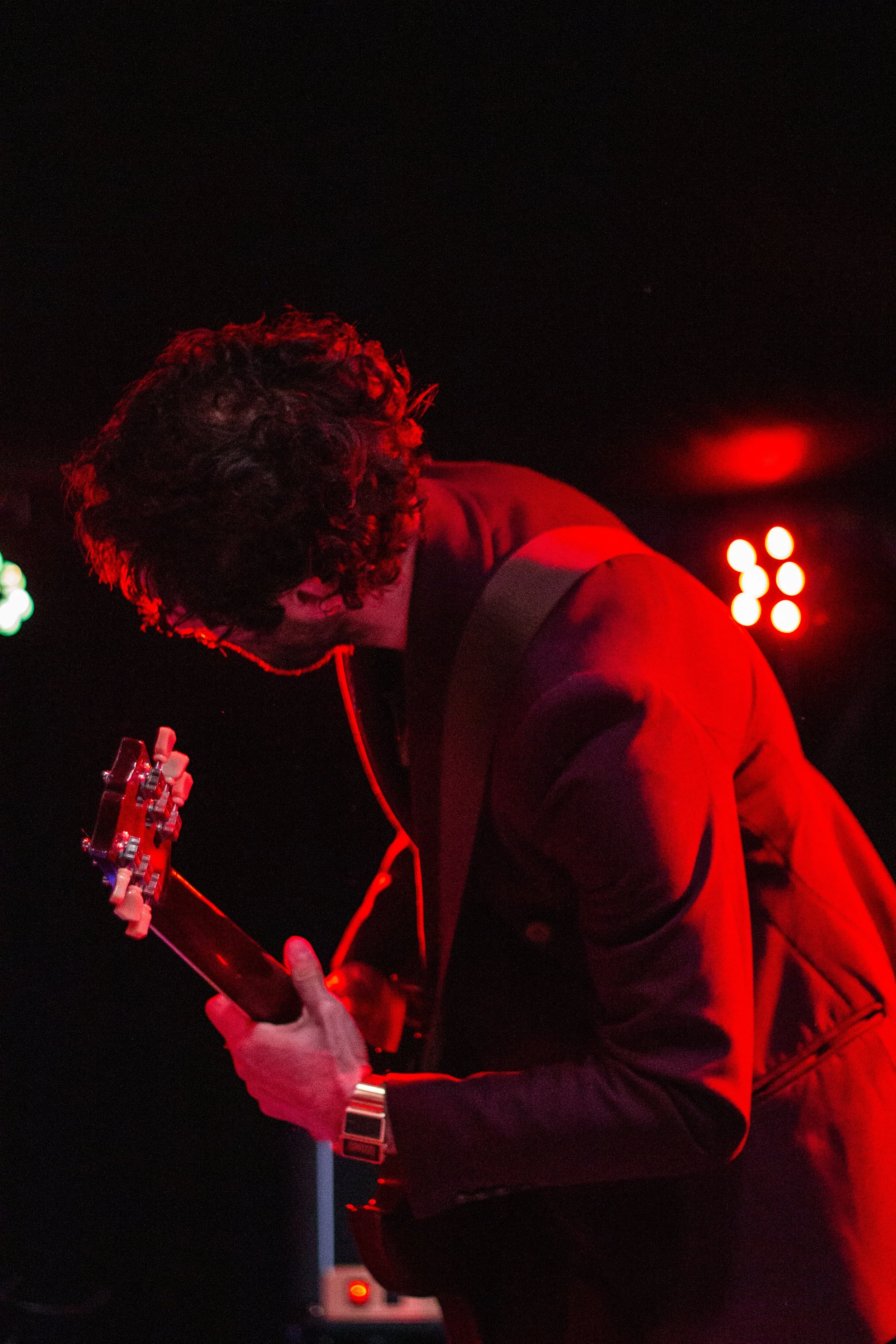 A person with curly hair playing an electric guitar, lit by red stage lights, wearing a dark jacket.