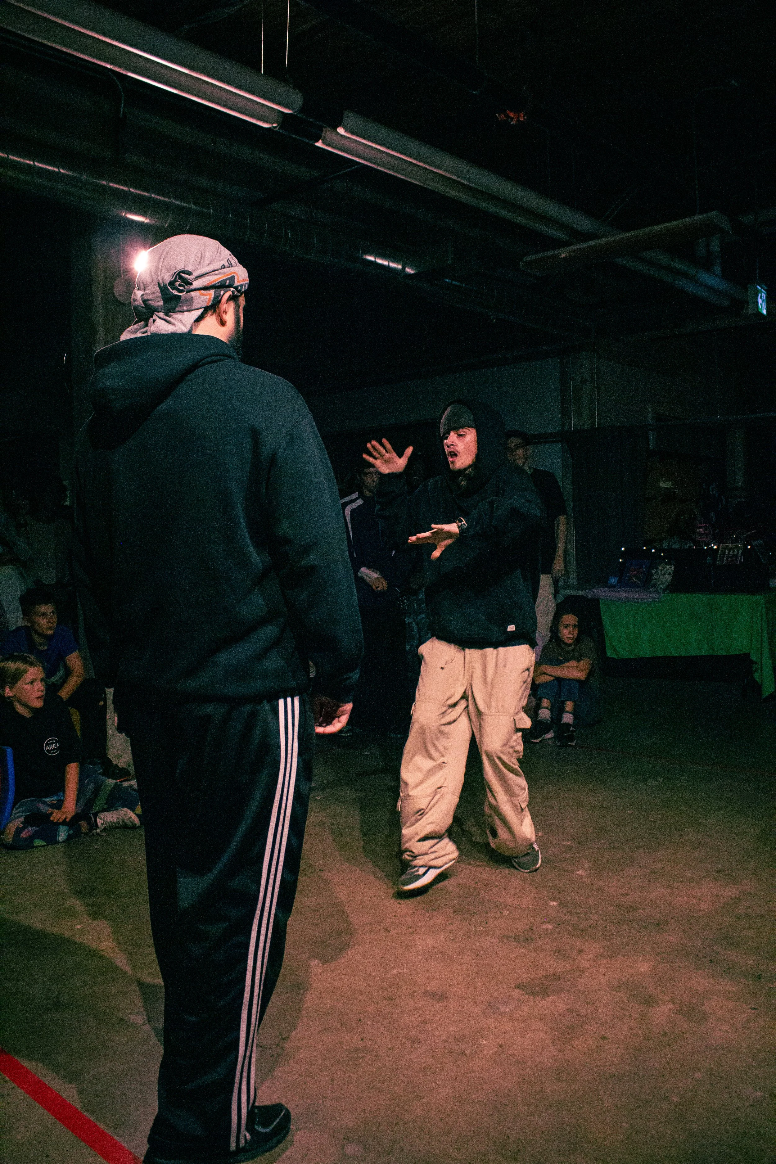 Two young men are in the middle of a dance battle in a dark indoor venue. One is wearing a gray headscarf and a black hoodie, and the other is wearing a black hoodie and beige cargo pants. Several people are watching in the background, some sitting o