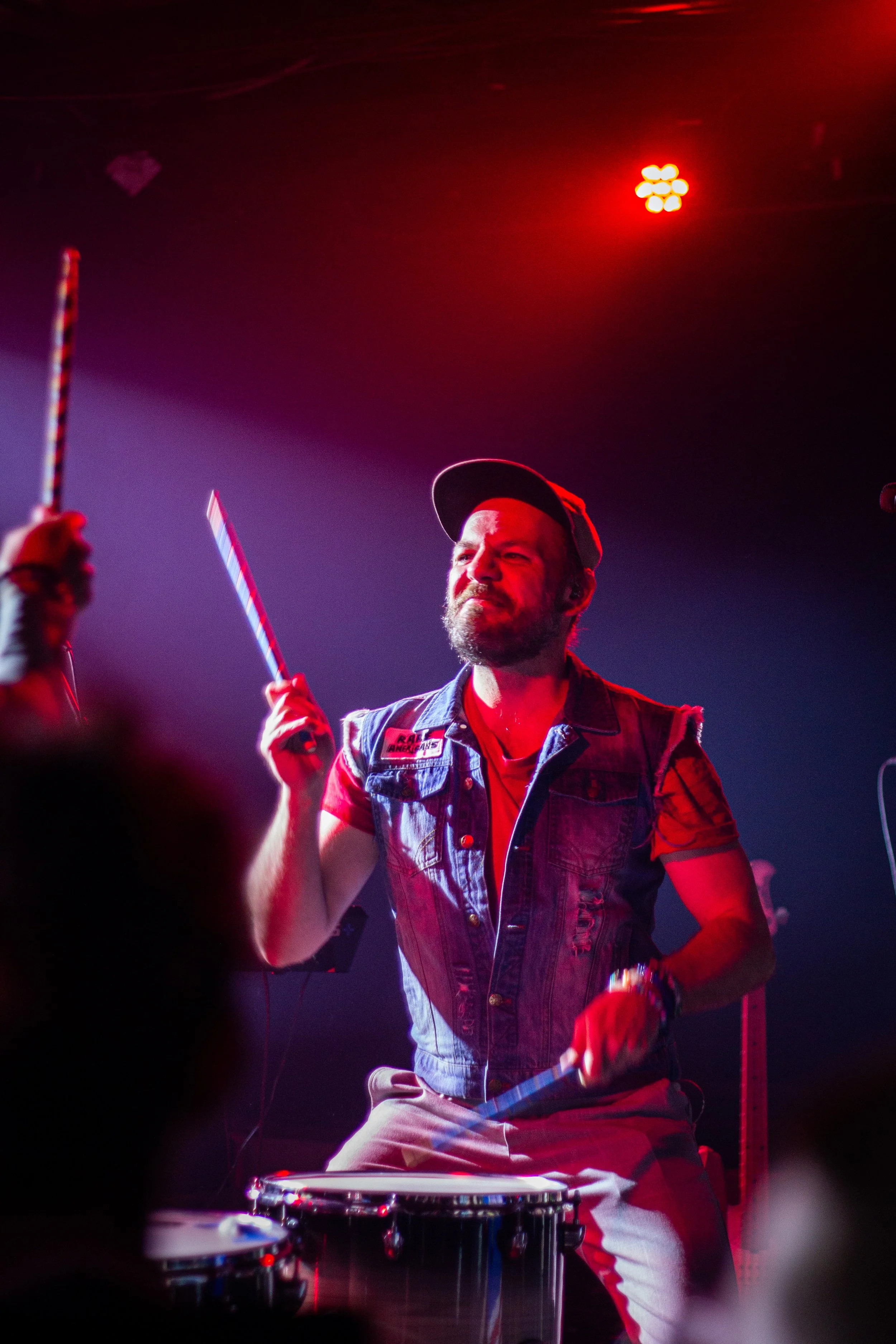 A man playing drums on stage, wearing a denim vest, baseball cap, and headphones, with stage red and purple lighting.