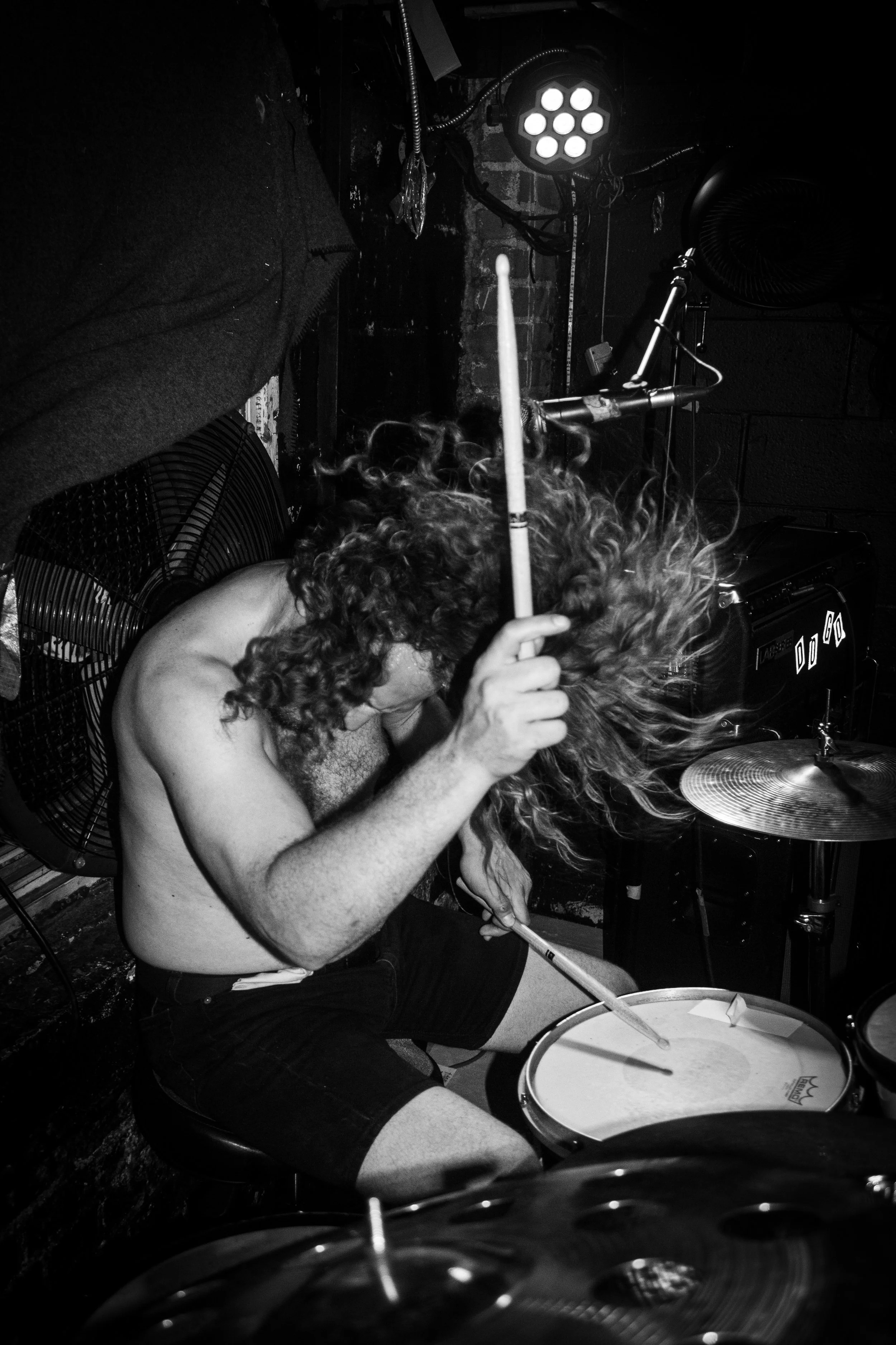 A shirtless man with curly hair playing drums in a dimly lit room with stage lighting and fans.