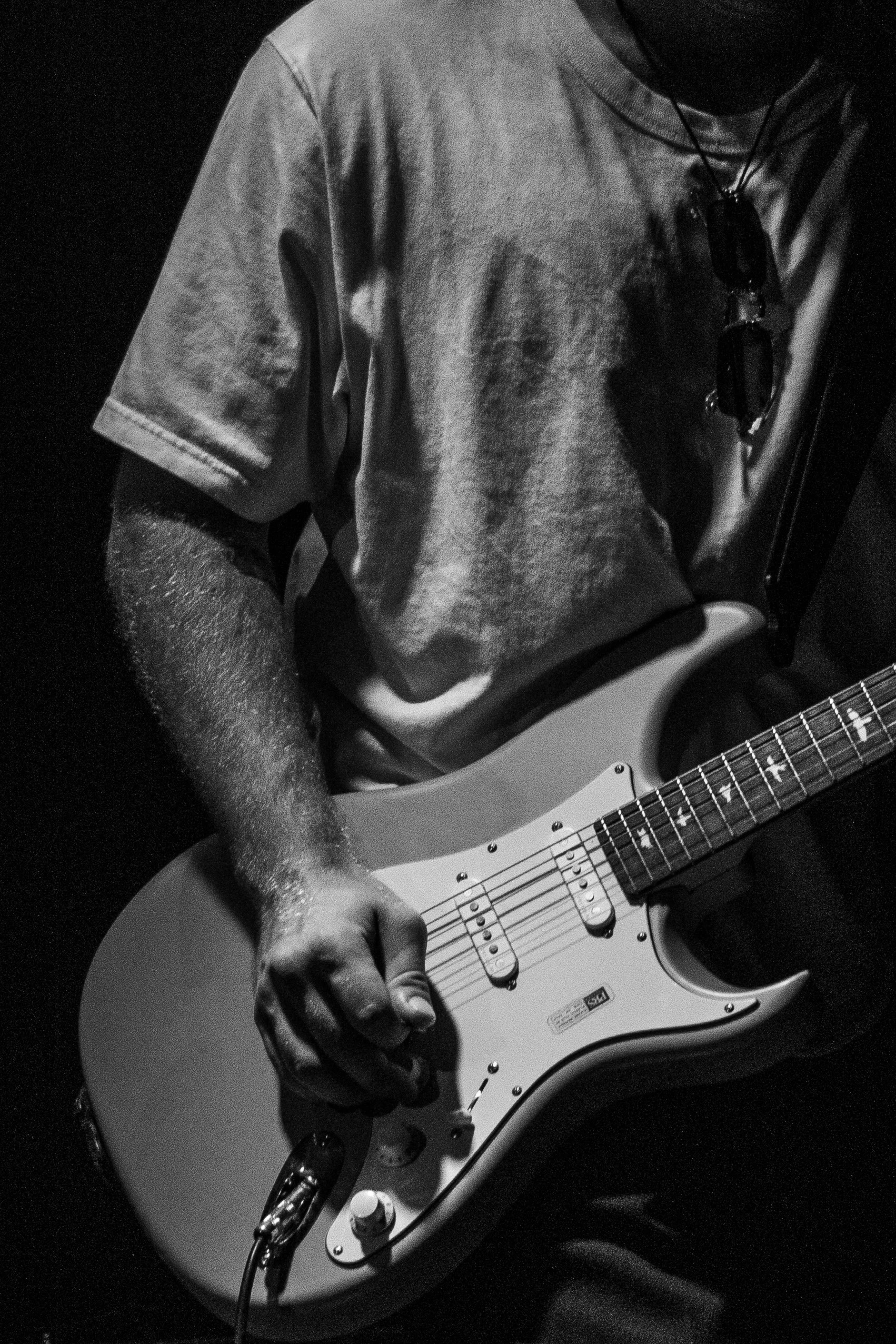 Person wearing a t-shirt playing an electric guitar, with focus on the guitar and hand, in a low-light setting.