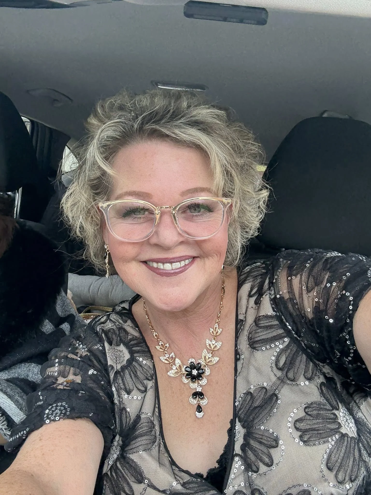 A woman with short curly blonde hair, wearing glasses, smiling in a car. She is dressed in a black and beige floral top with lace accents, gold jewelry including earrings, a necklace with black and white flower pendant, and makeup with lipstick.