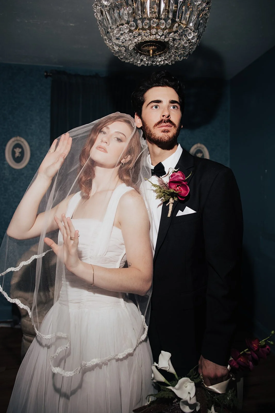 A bride and groom pose indoors for a wedding photo. The bride is wearing a white dress and veil, with her hand near her face. The groom is dressed in a black tuxedo holding flowers. A chandelier hangs above them.
