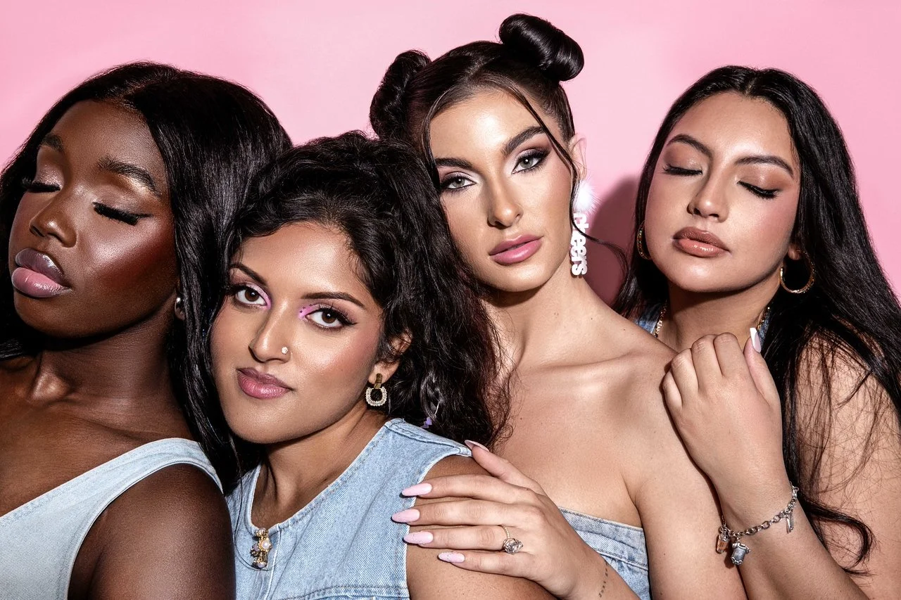 Four women with diverse skin tones and dark hair, posing closely together against a pink background, with some eyes closed and some looking at the camera.