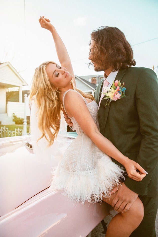 A woman in a white dress and a man in a dark suit with a boutonniere are dancing outdoors near a vintage car, with a house visible in the background.