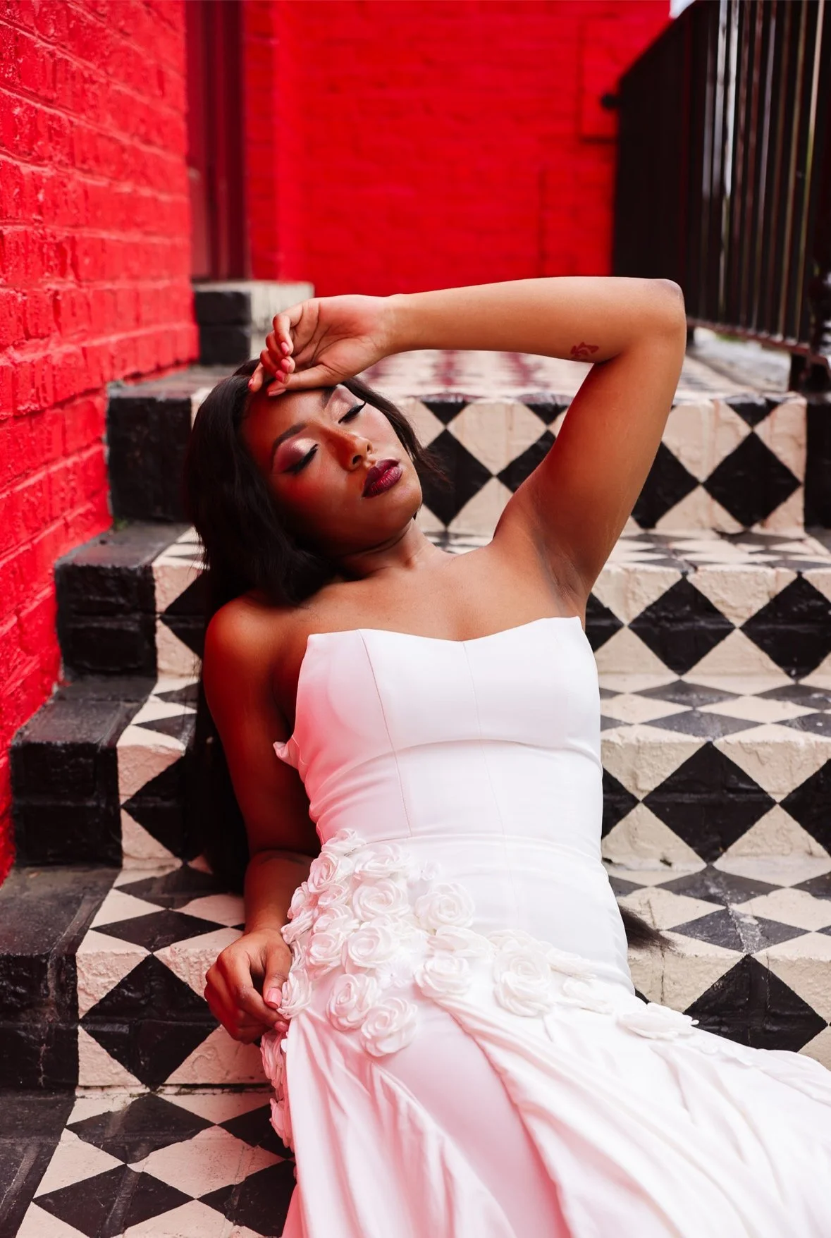 A woman in a white dress with floral details sitting on black and white patterned stairs against a red brick wall with a railing nearby.