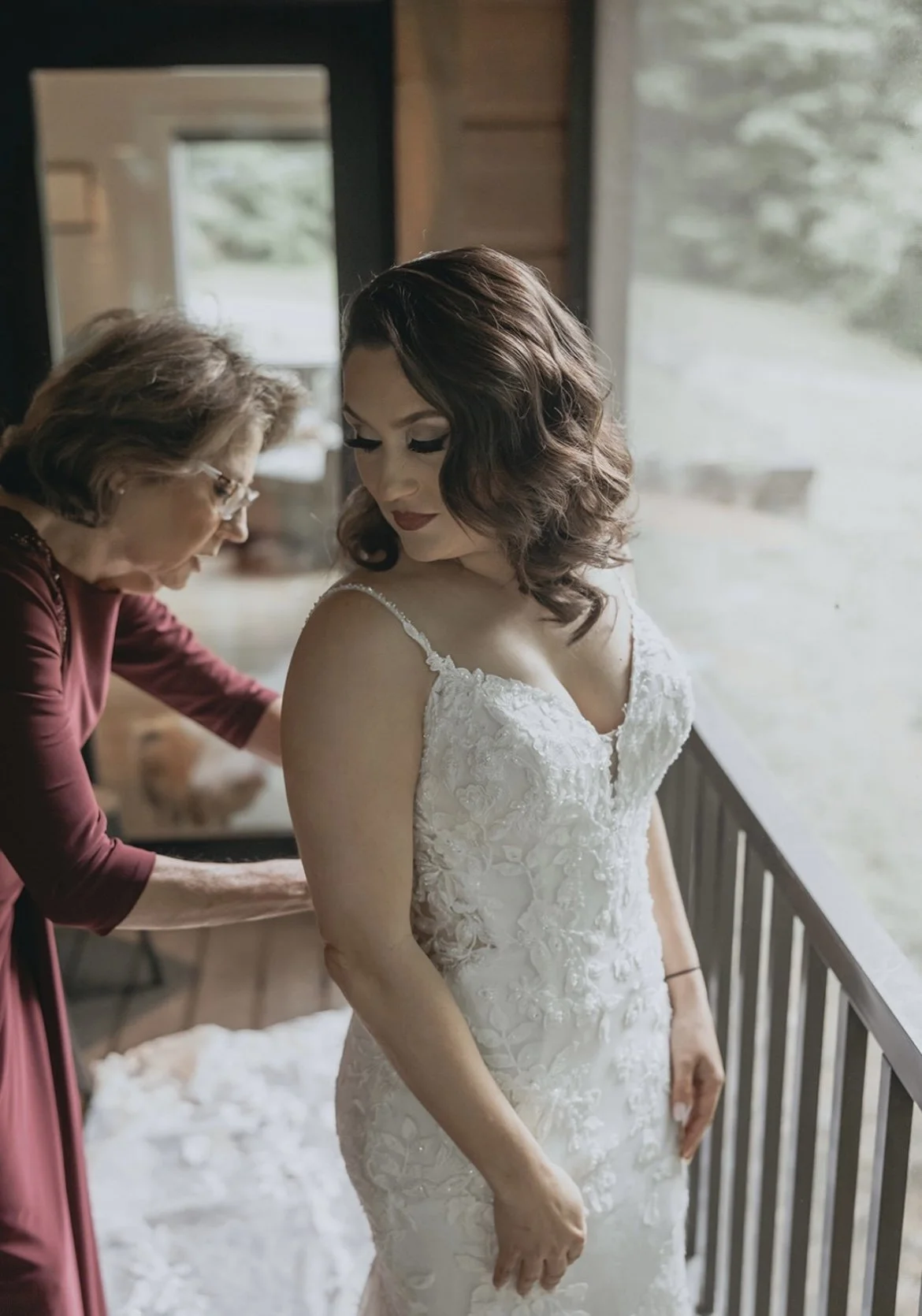 Bride in white wedding dress being assisted by older woman indoors.