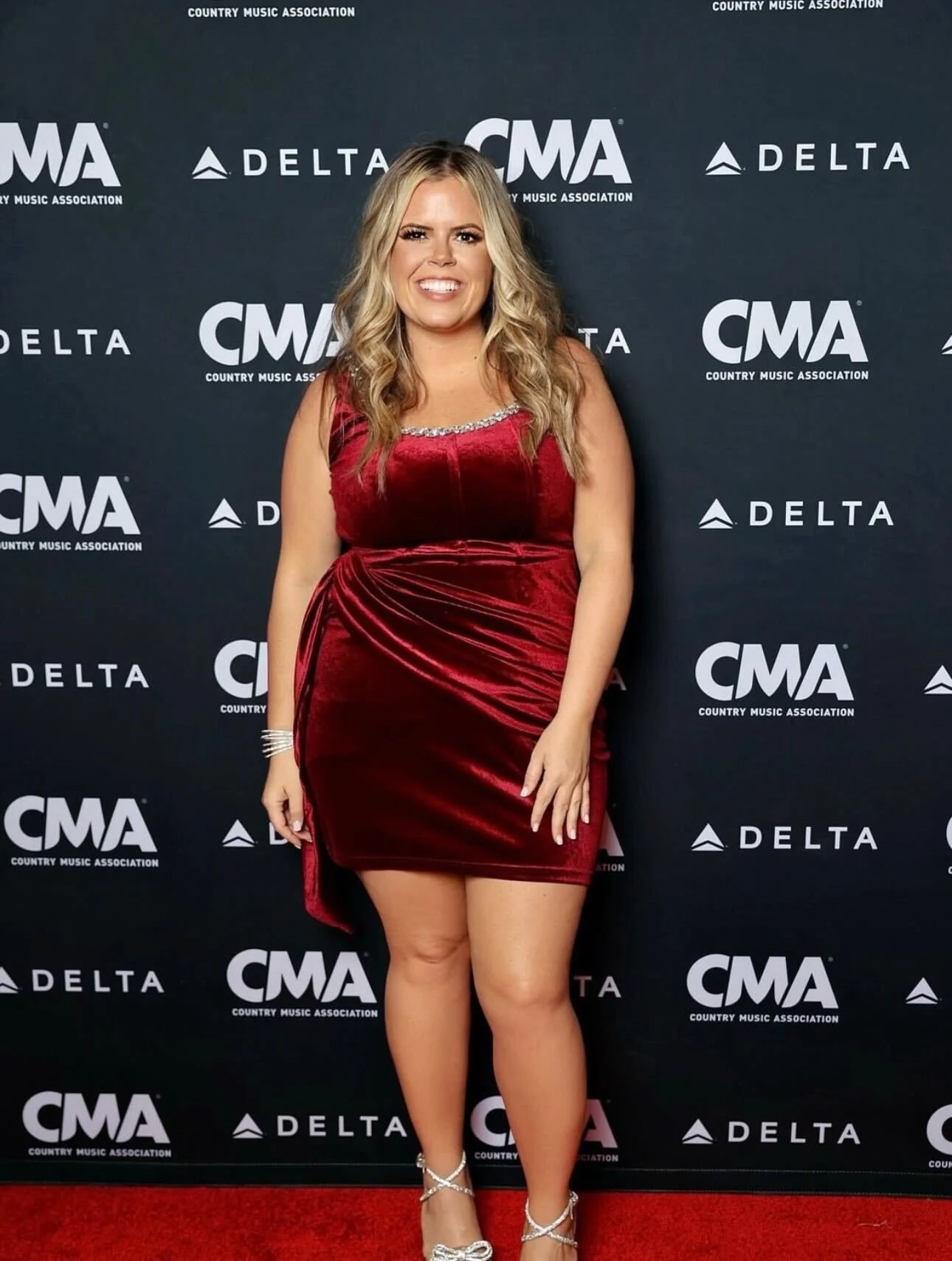 Woman wearing soft glam CMA makeup with radiant skin, defined eyes, and a neutral lip while posing in a red velvet dress on the CMA Awards step-and-repeat backdrop.