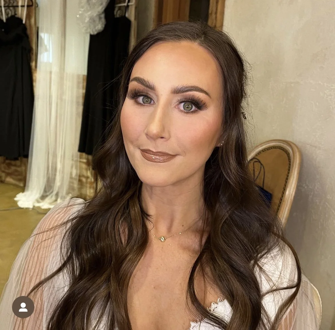 Woman with long brown hair and makeup, wearing a white top, standing indoors with black dresses in the background.