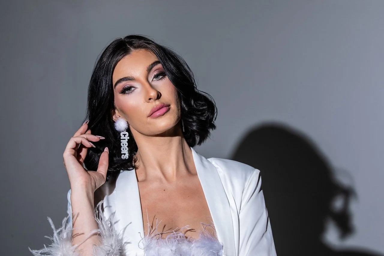 A woman with dark, wavy hair winks at the camera, wearing a white satin robe with feathered cuffs, pink lipstick, and statement earrings that spell 'glam' with a fuzzy pom-pom.
