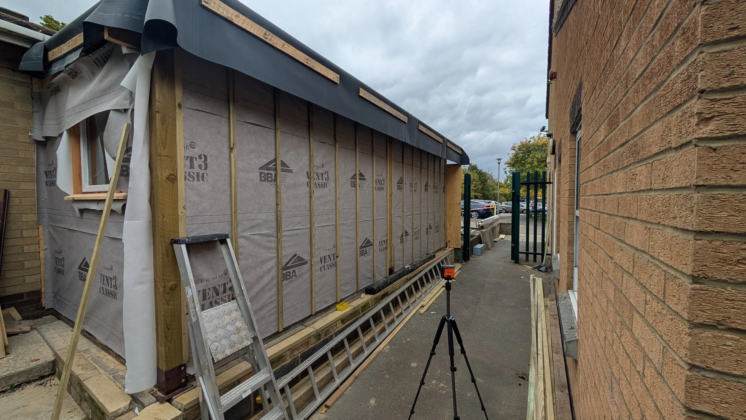 Exterior of a building under construction with framing and sheathing, a ladder leaning against the wall, and a work light on a tripod in the sidewalk.