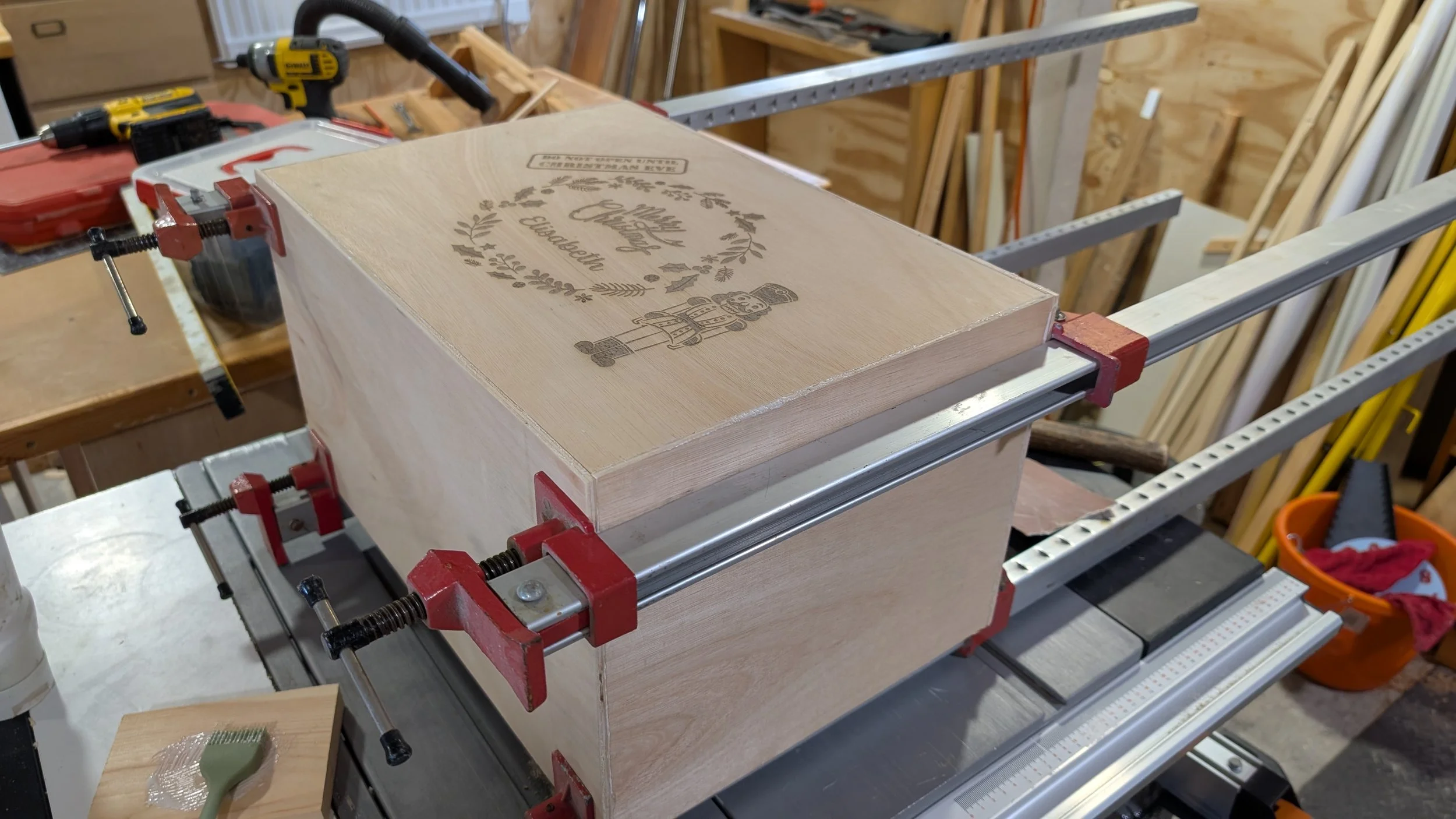 Wooden box clamped to a workbench in a woodworking shop with engraved text and design on the lid.
