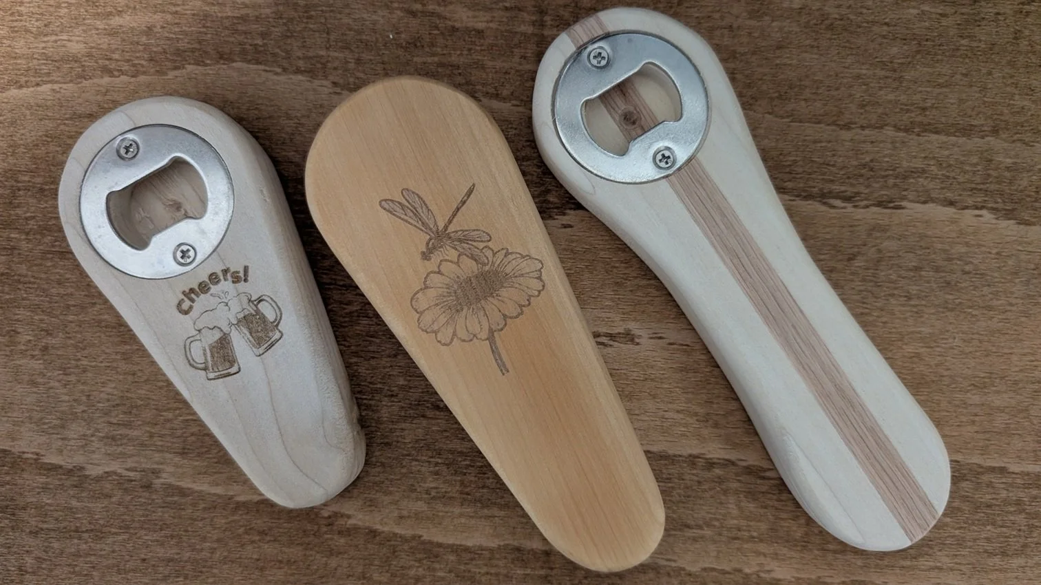 Three outdoor corkscrews with engraved designs, one with beer mugs and 'Cheers!' text, one with a dragonfly and flower, and one with a stripe pattern, all placed on a wooden surface.