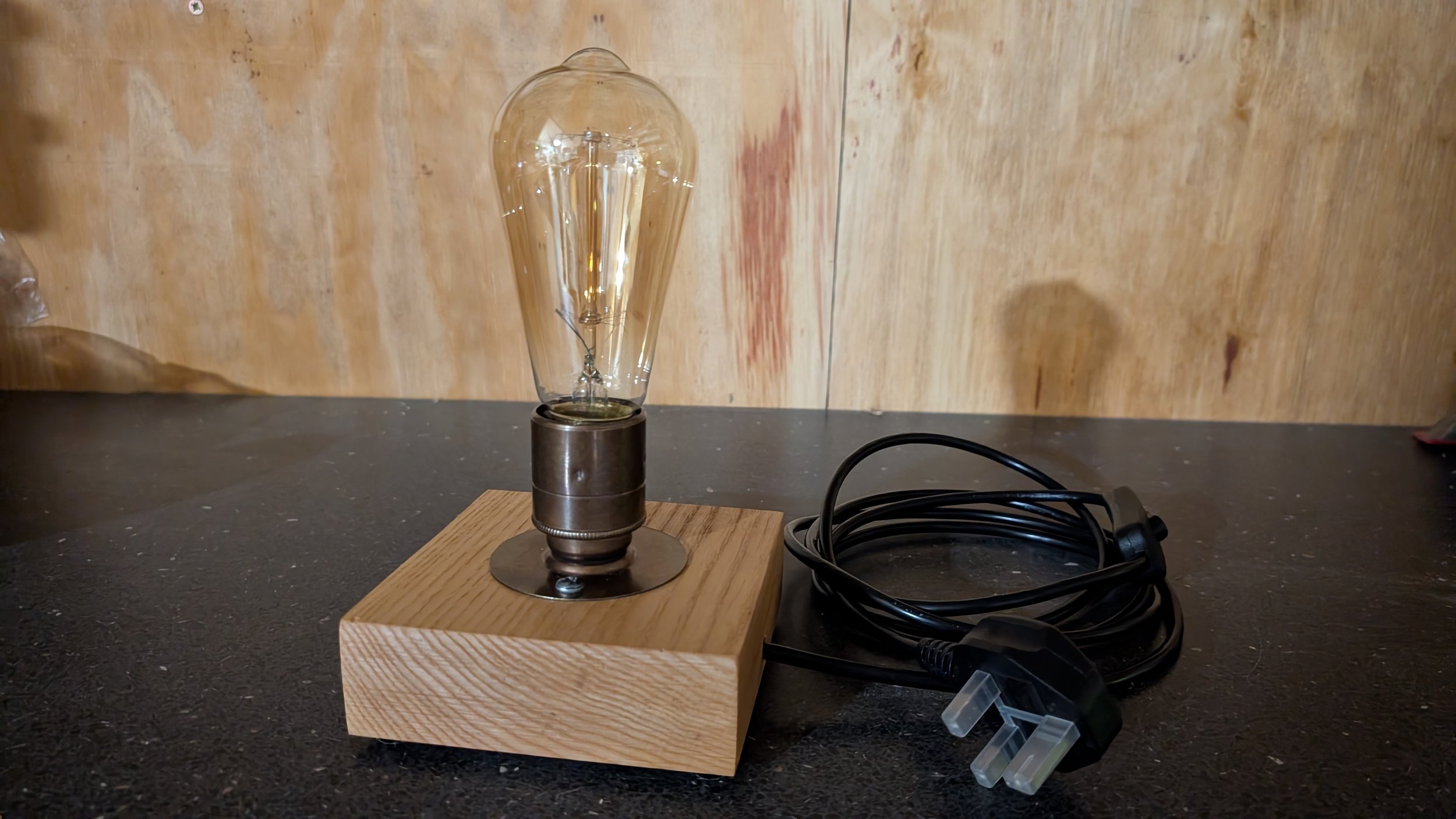 A vintage-style light bulb mounted on a wooden base with a black power cord coiled nearby on a black surface against a wood-paneled background.