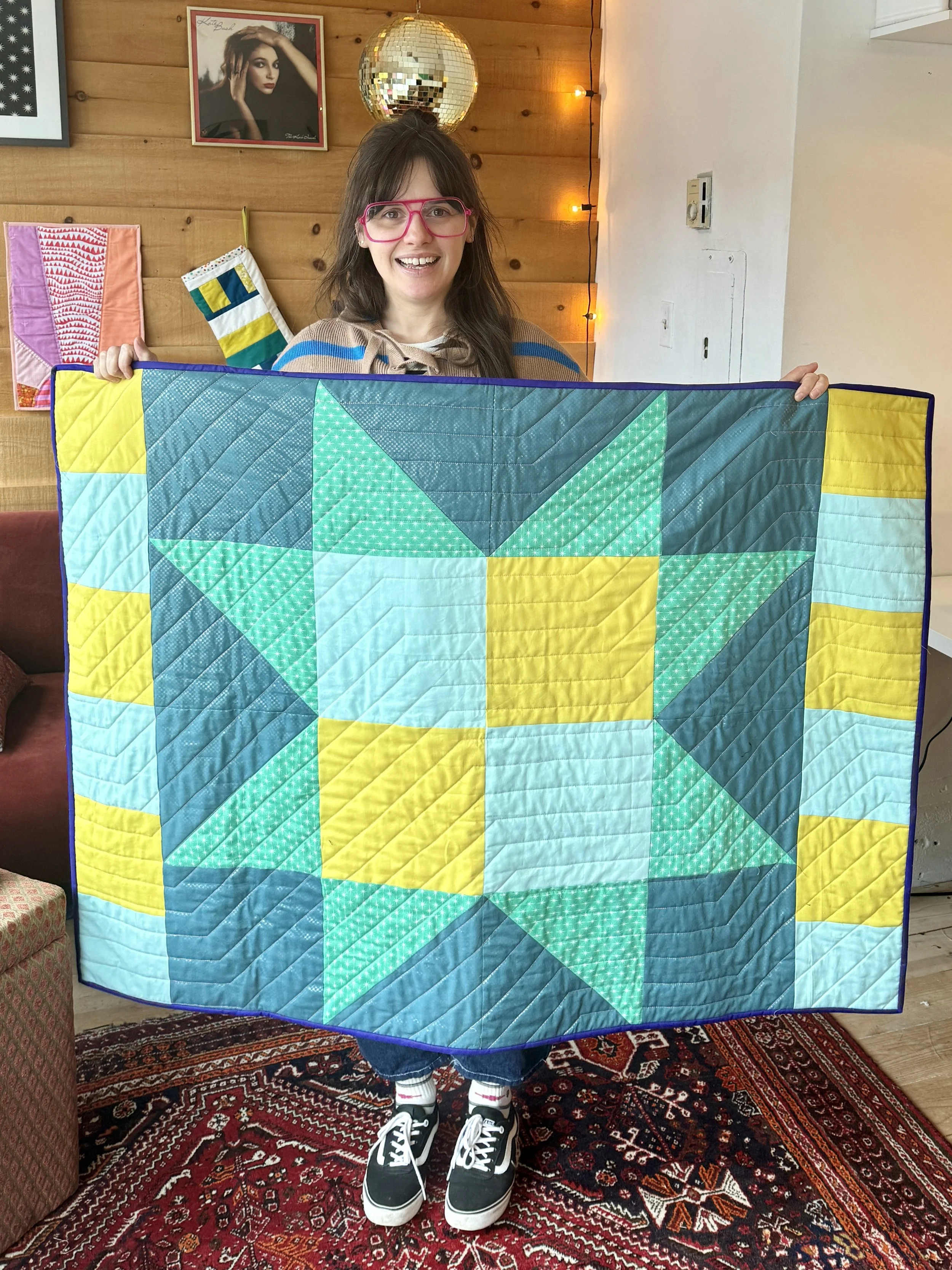 Alex is holding up her quilt that she made. It is a sawtooth star lap quilt