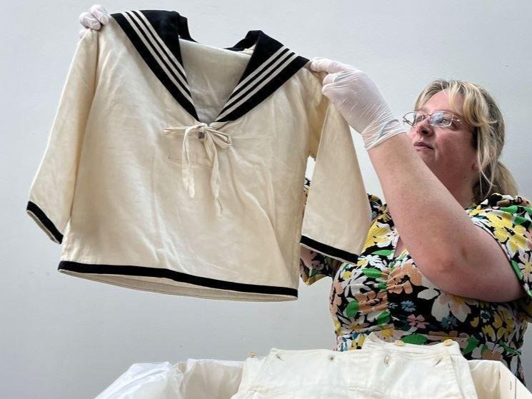 A woman with gloved hands holds up the artifact: he striped sailor suit that Prince Albert wore in 1846, to board the Royal Yacht. This outfit sparked a massive trend.