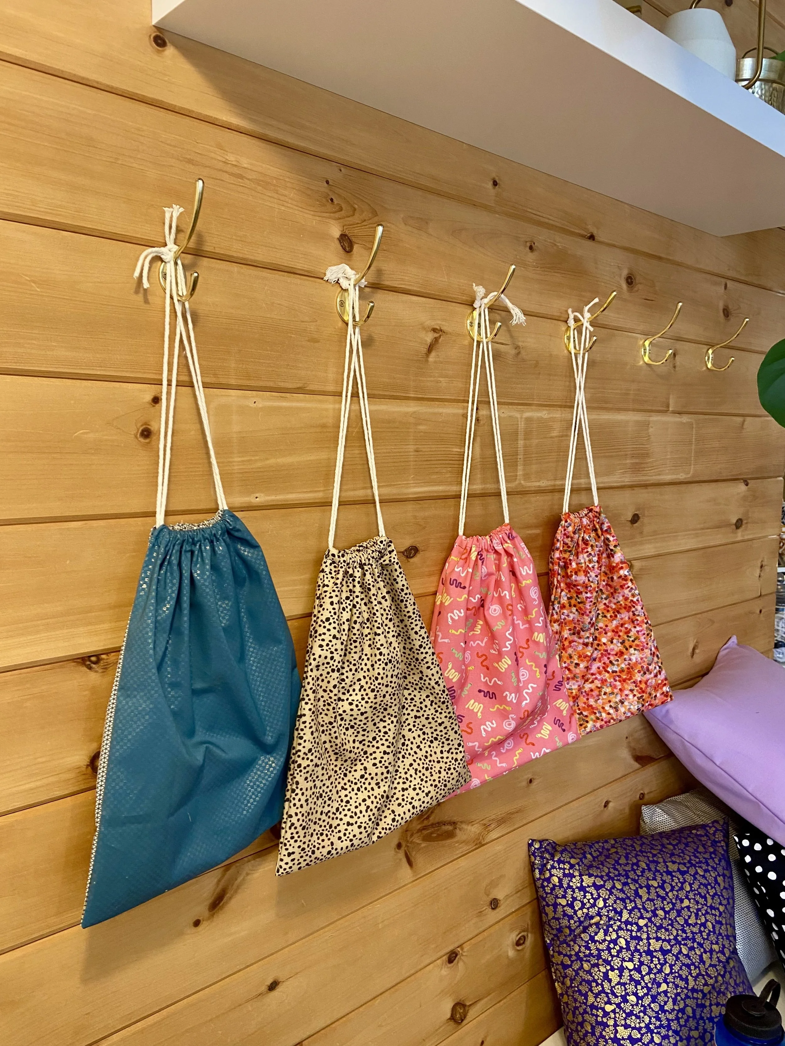 Four colorful fabric drawstring bags hanging from gold hooks on a wooden wall, with pillows and other items nearby.