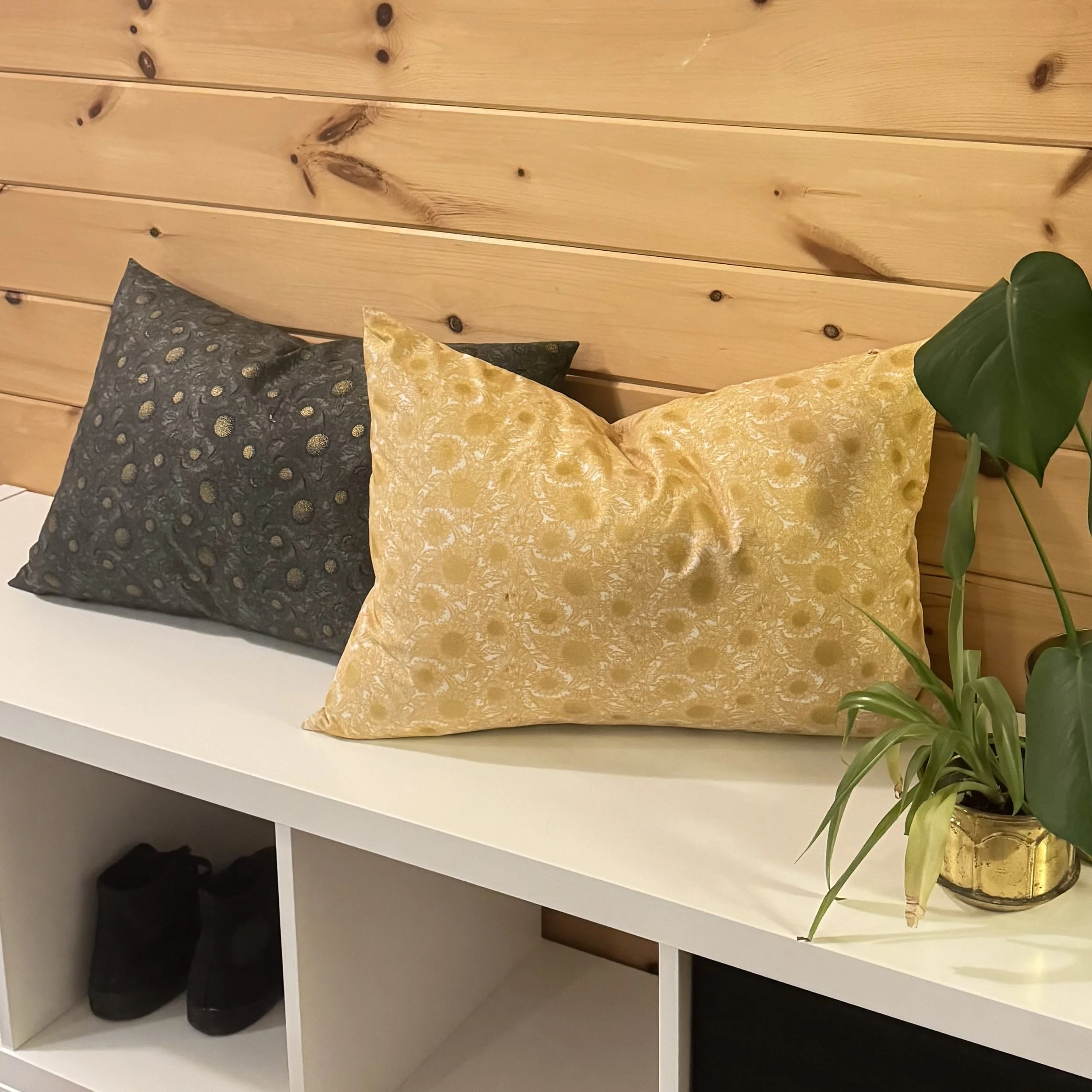Two decorative pillows, one black with gold polka dots and one gold with a floral pattern, are on a white shelf against a wooden wall. A potted plant with large green leaves is on the right side of the pillows.