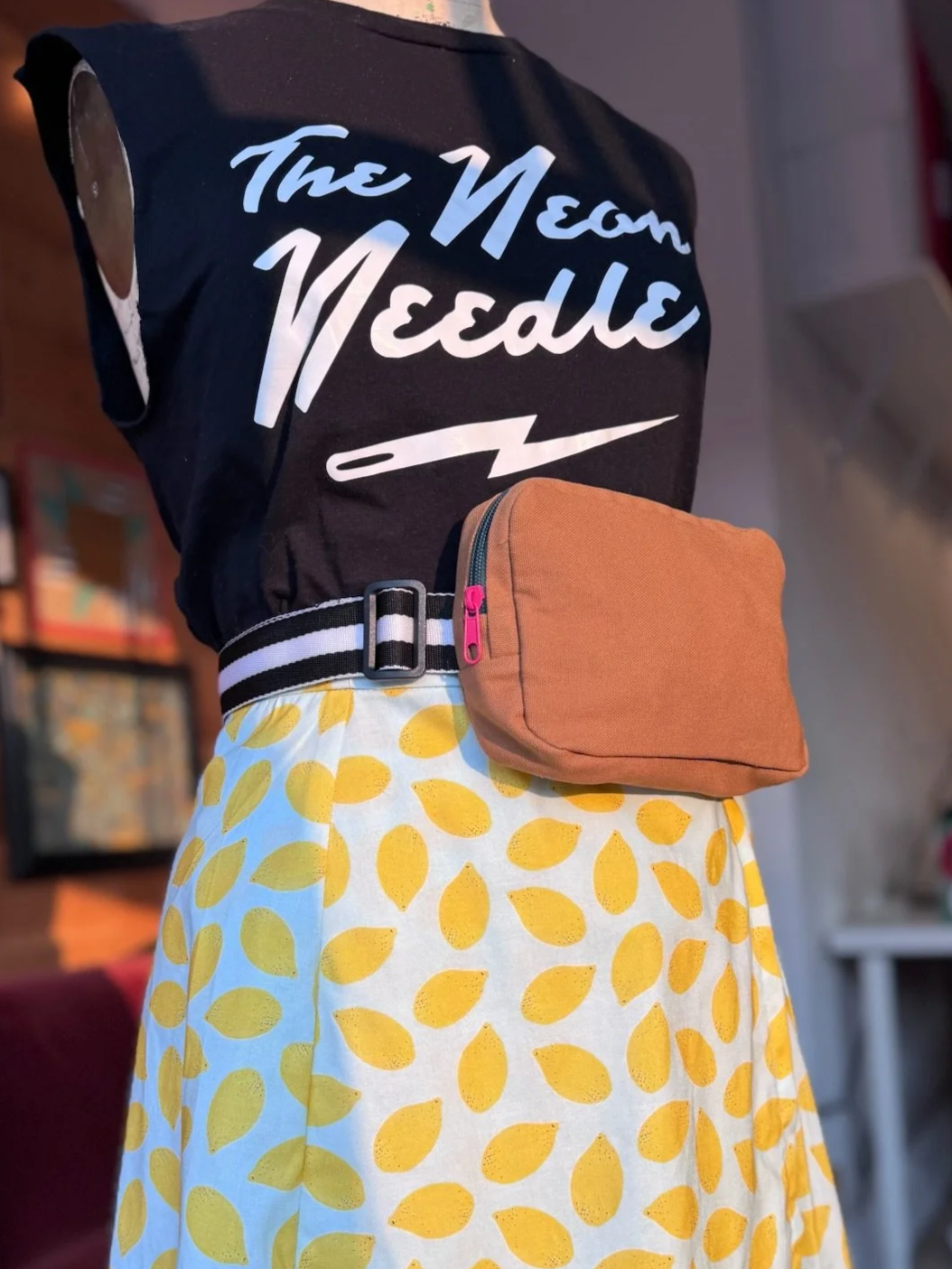 Close-up of a mannequin wearing a black sleeveless shirt with white script that says 'The Nehem' and yellow and white skirt with a lemon pattern, a black and white striped belt, and a brown waist bag. Blurred indoor background.