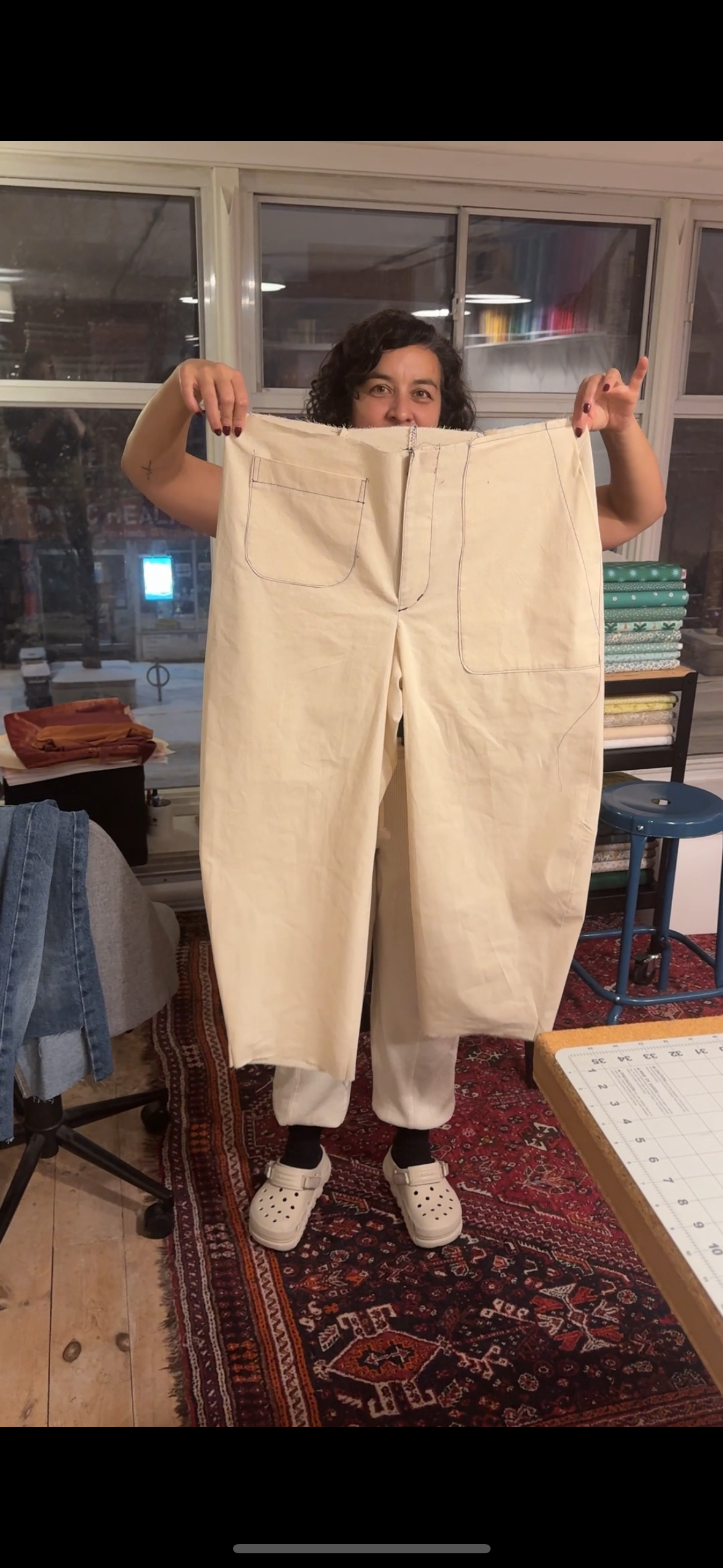 A woman standing indoors holding up a large pair of cream-colored pants with visible pocket and seam details, in front of a window with shelves and fabric rolls in the background.