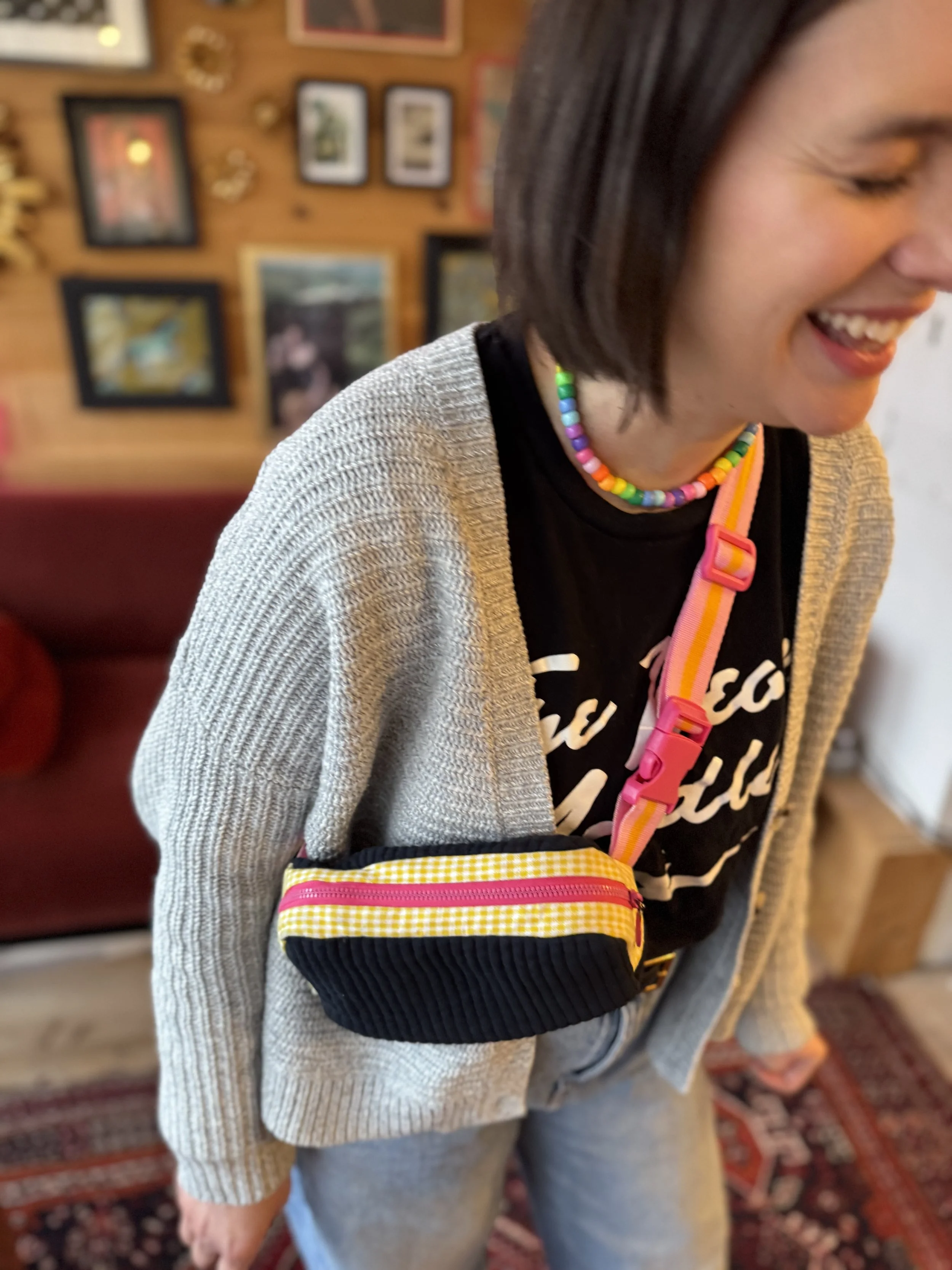 A woman smiling, wearing a rainbow beaded necklace, a black T-shirt, a beige cardigan, and a pink and yellow crossbody bag.