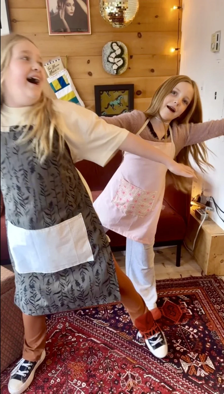 Two girls having fun in a cozy, decorated room with wooden walls. One girl is wearing a patterned apron and is extending her arm. The other girl is wearing a pink apron and beige pants, with arms wide open. The room has framed pictures, a disco ball,