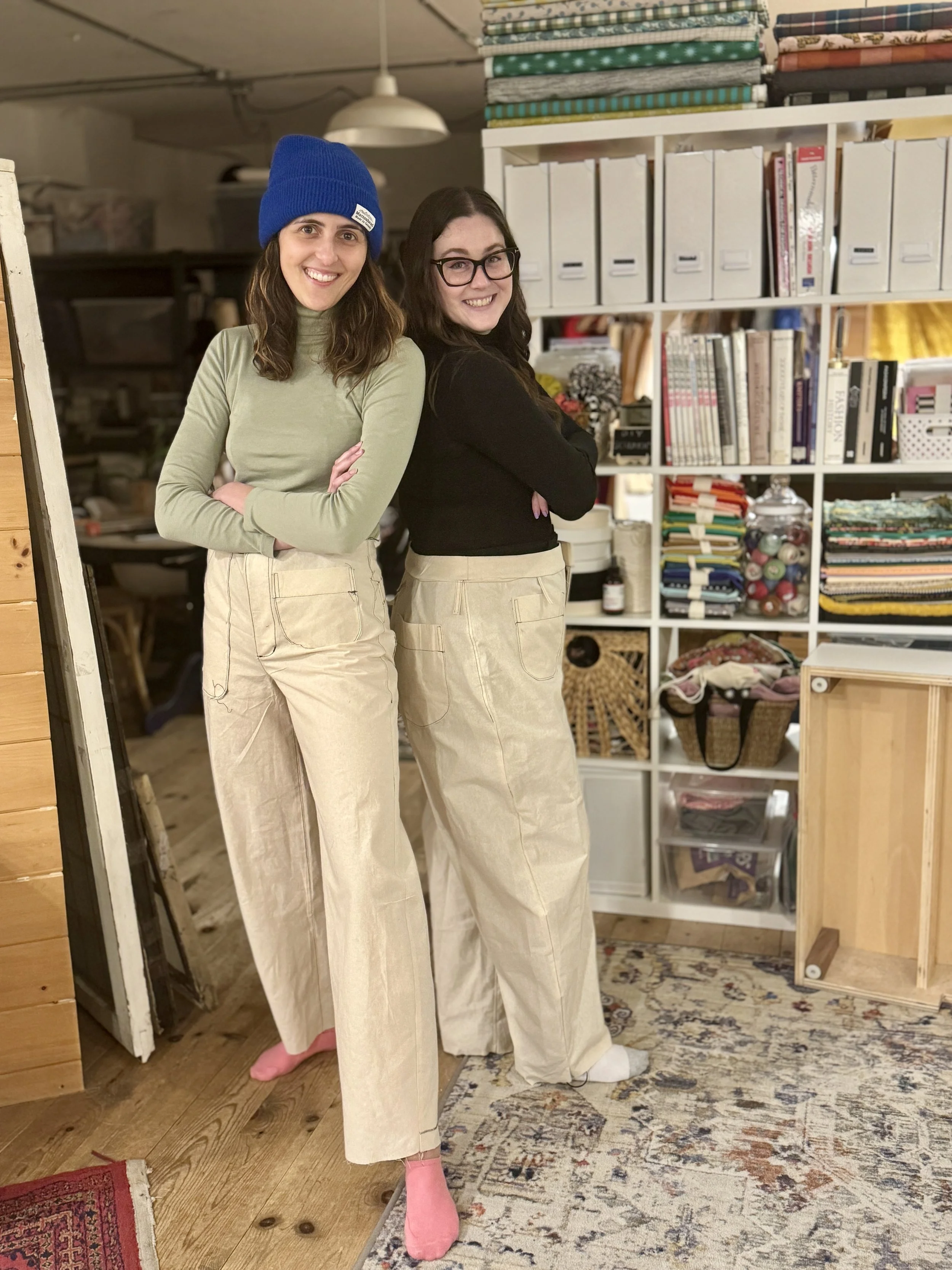 Two women standing indoors in front of a shelving unit filled with books, papers, and sewing supplies, smiling with arms crossed, one wearing a blue beanie and a light green turtleneck, the other wearing glasses and a black top.