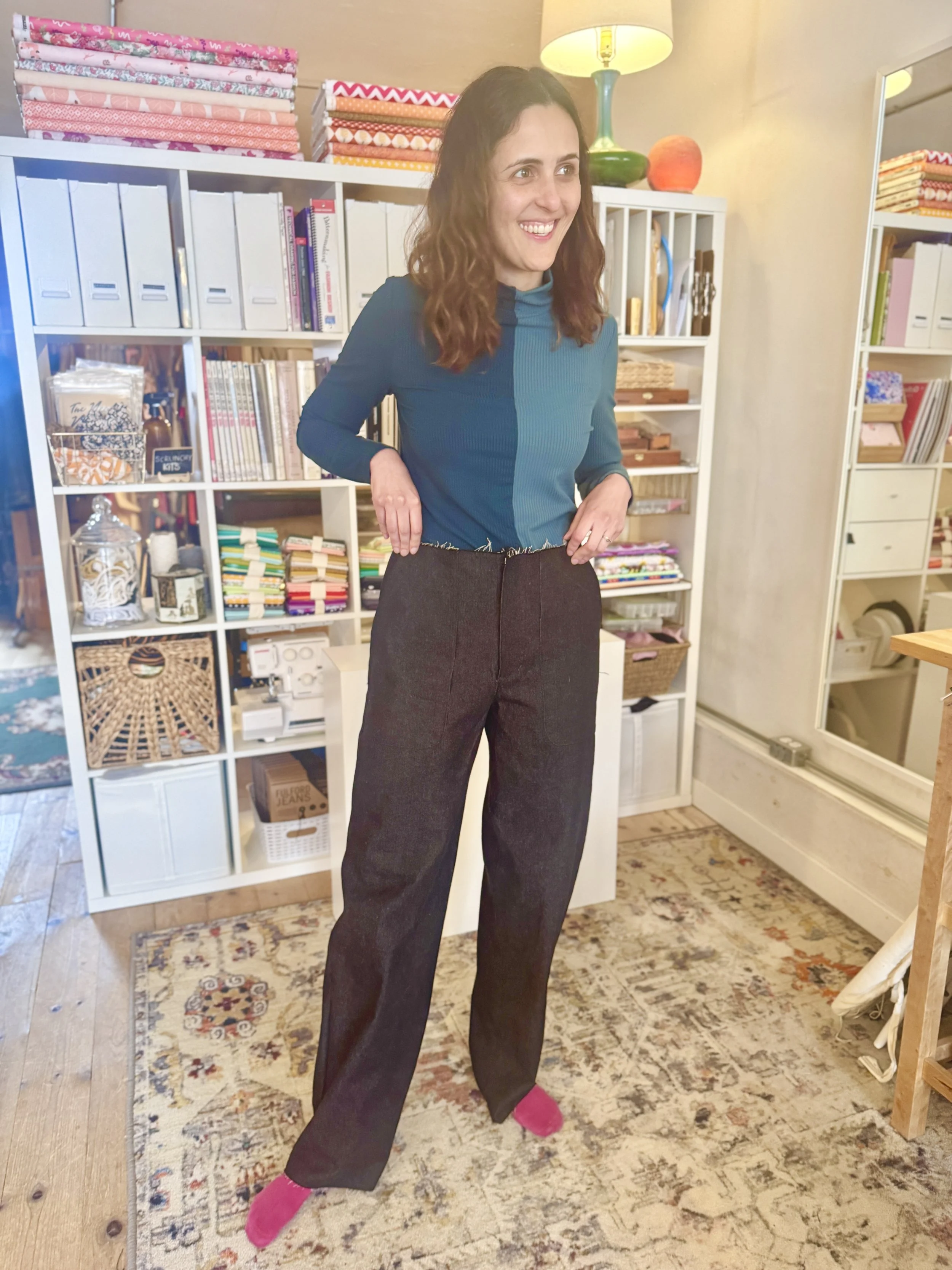 A woman with brown, wavy hair wearing a teal long-sleeve top and dark trousers standing in a craft or fabric store, smiling, with shelves of fabric and craft supplies behind her.
