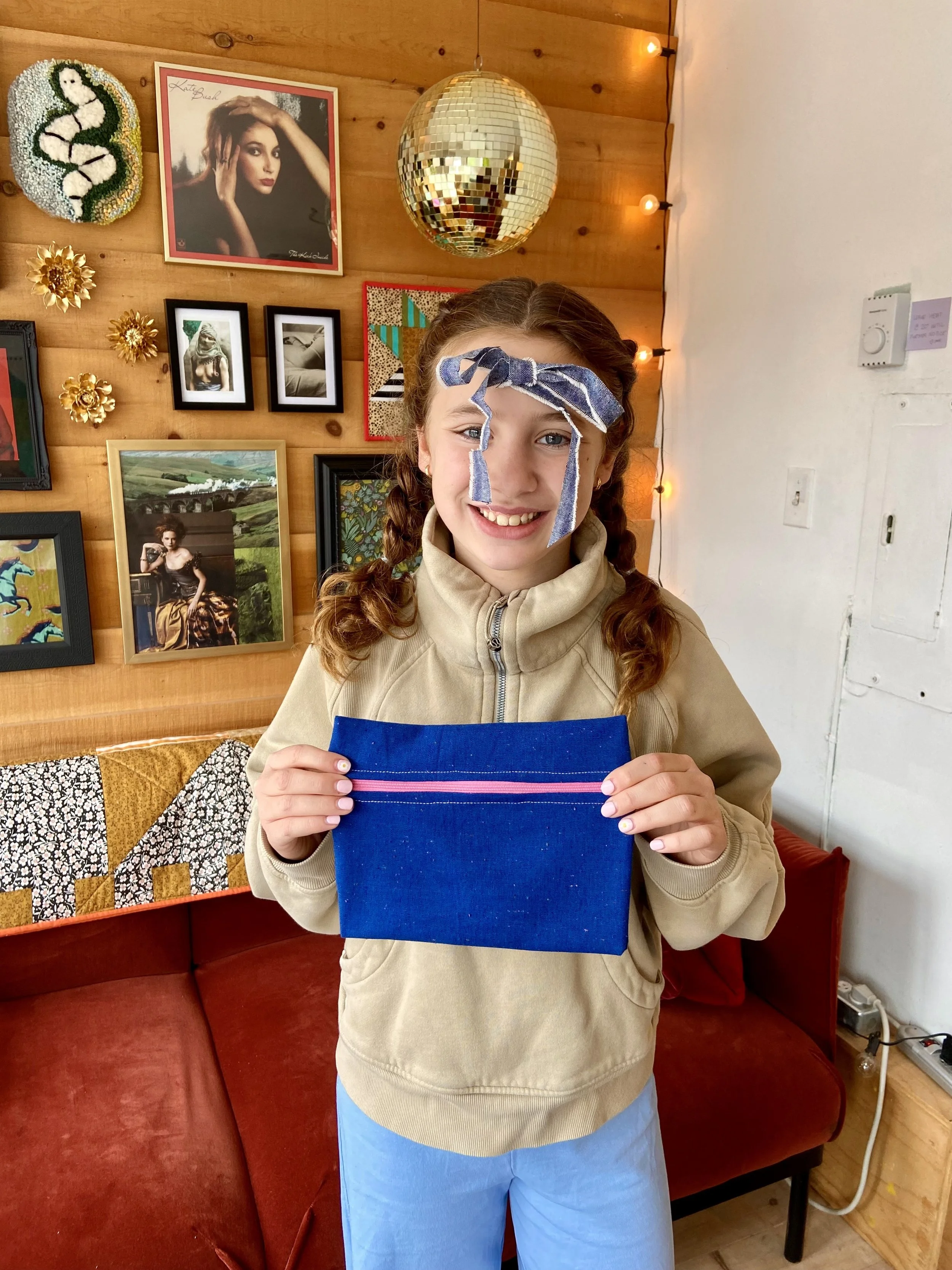 A girl with braided hair and a fabric bow on her head, smiling and holding a blue zippered pouch, standing indoors with a wall decorated with framed photographs, artwork, gold floral decorations, and a disco ball.