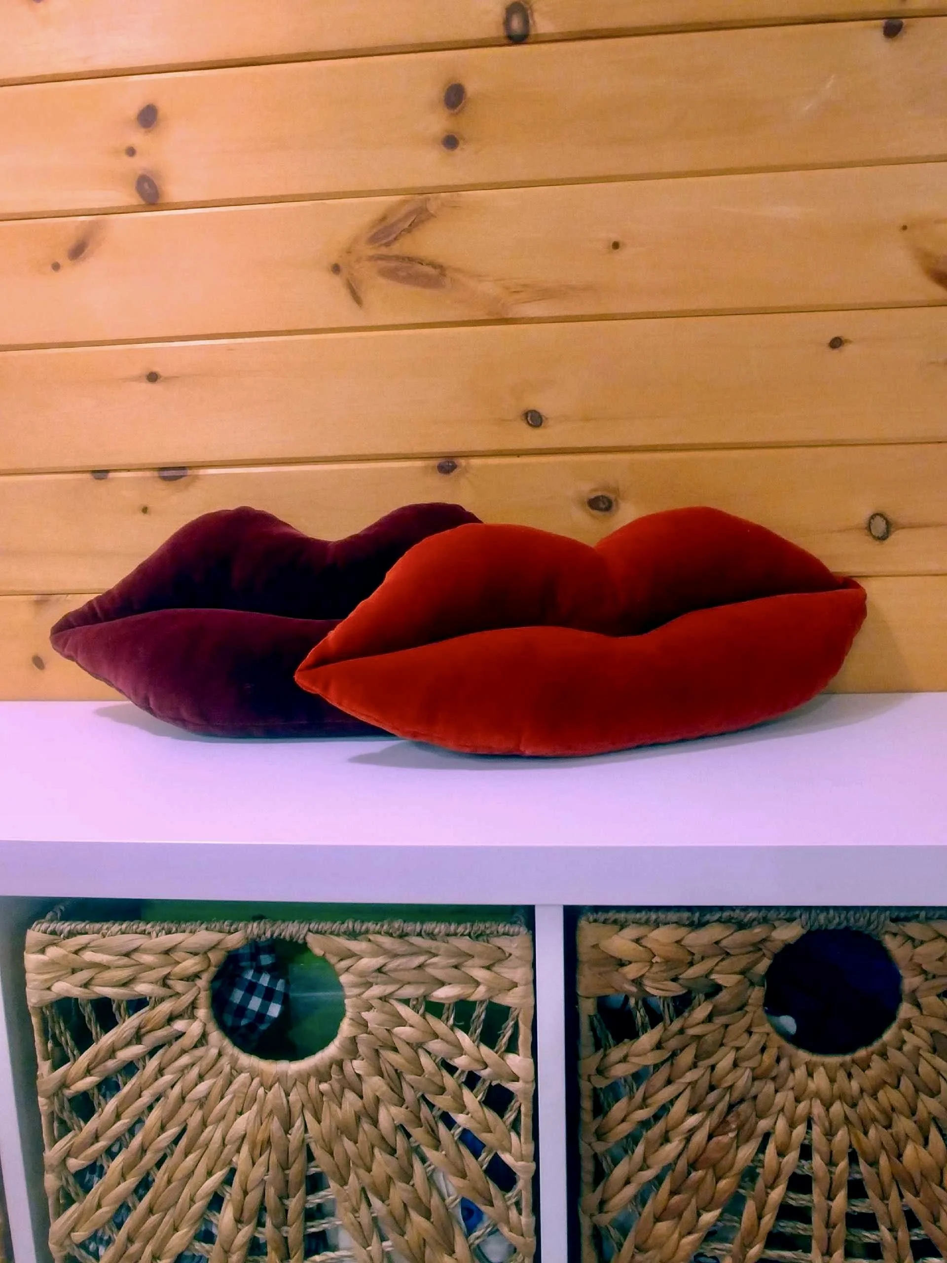 Two fabric pillows shaped like lips, one red and one dark purple, placed on a white shelf against a wooden panel wall.