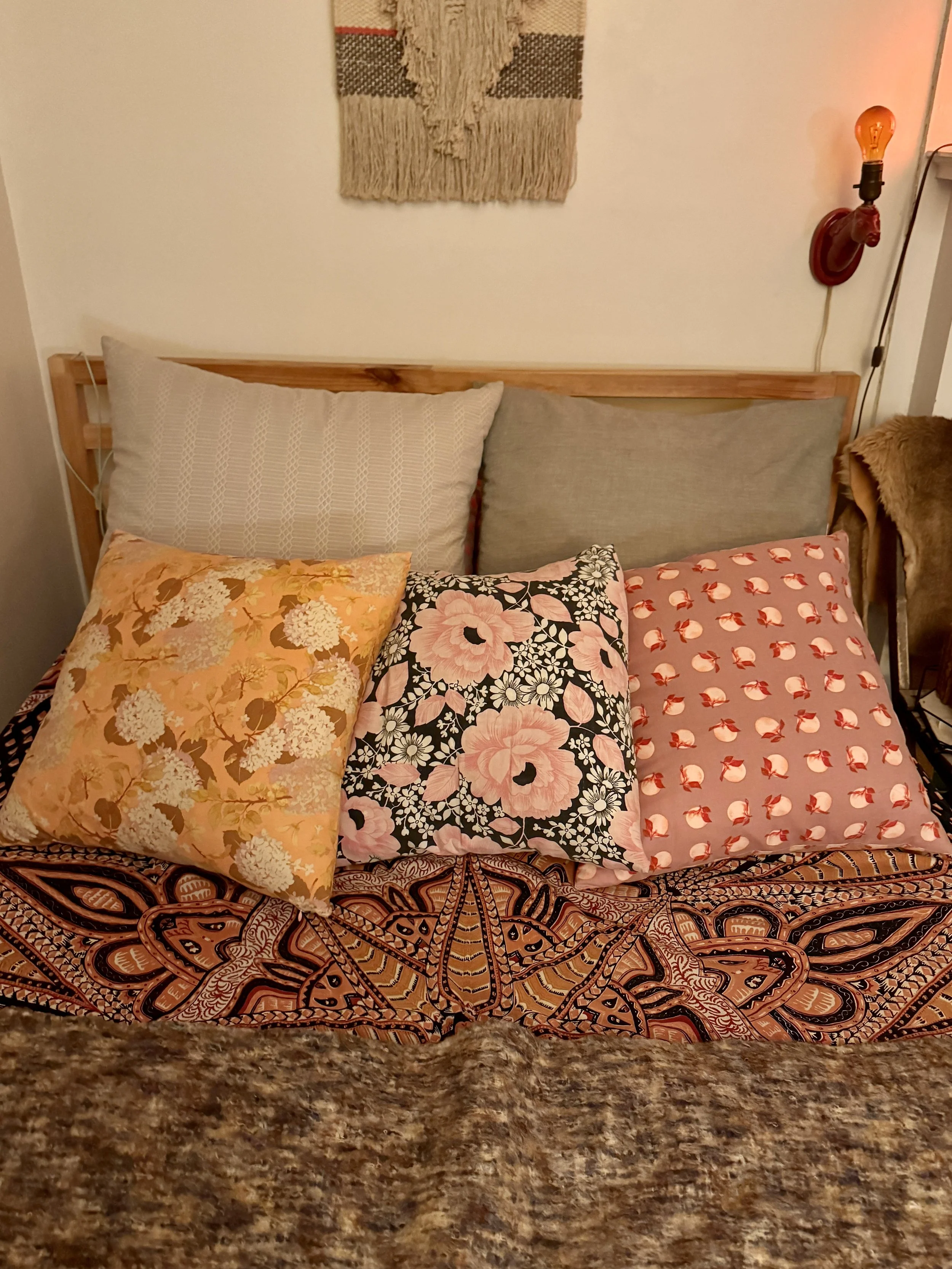 A bed with a colorful, patterned bedspread and three decorative pillows, against a plain wall with a woven wall hanging and a red wall-mounted light fixture.