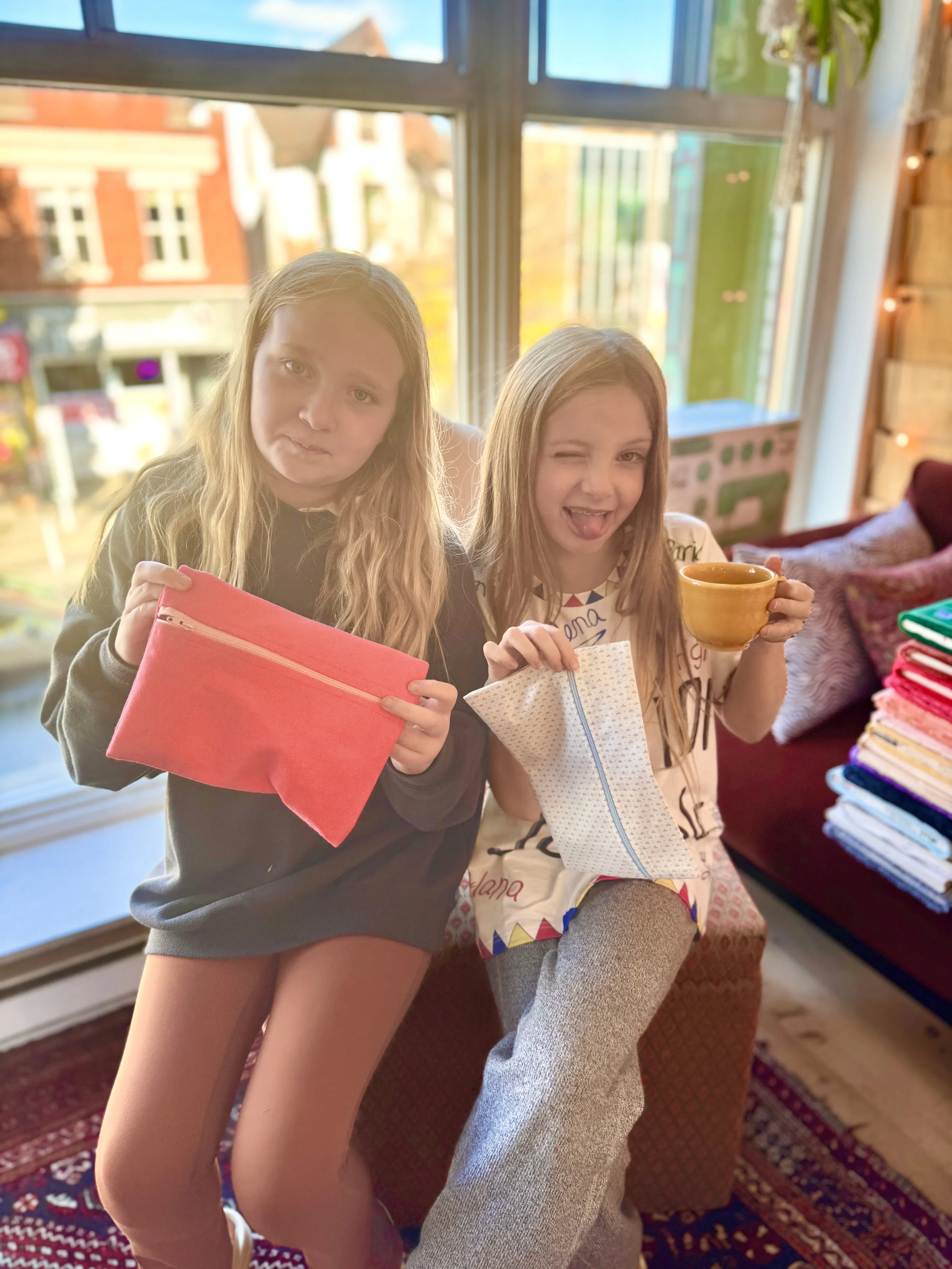 Two young girls sitting on a cushioned bench indoors near a large window. One girl is holding a pink pouch, and the other girl is holding a yellow mug and a white pouch with a blue zipper. They are smiling and making playful faces, with a colorful bl