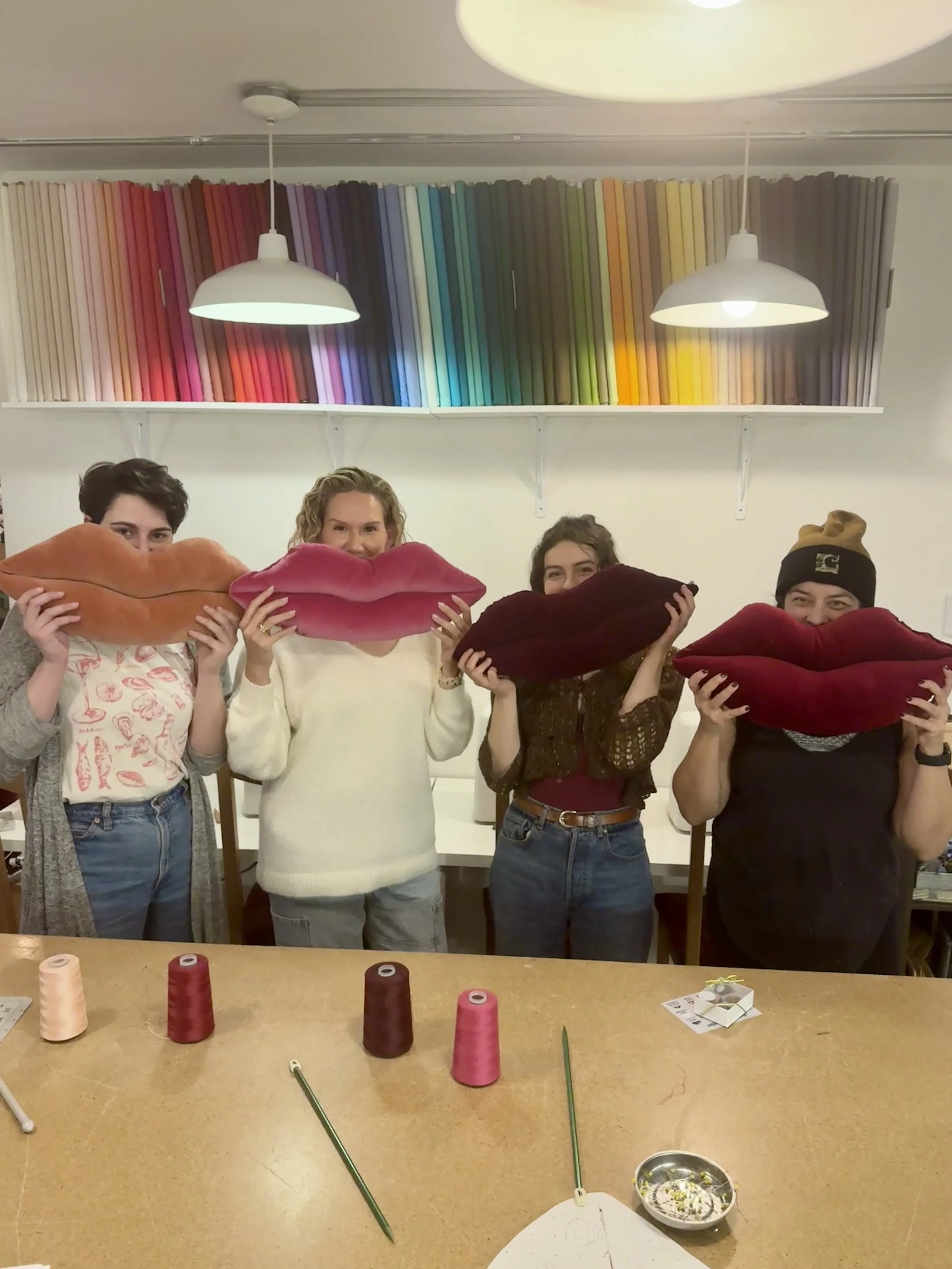 Four people standing behind a table, each holding a large lip-shaped pillow in front of their faces, in a room with shelves of colorful fabric swatches and two hanging lights.