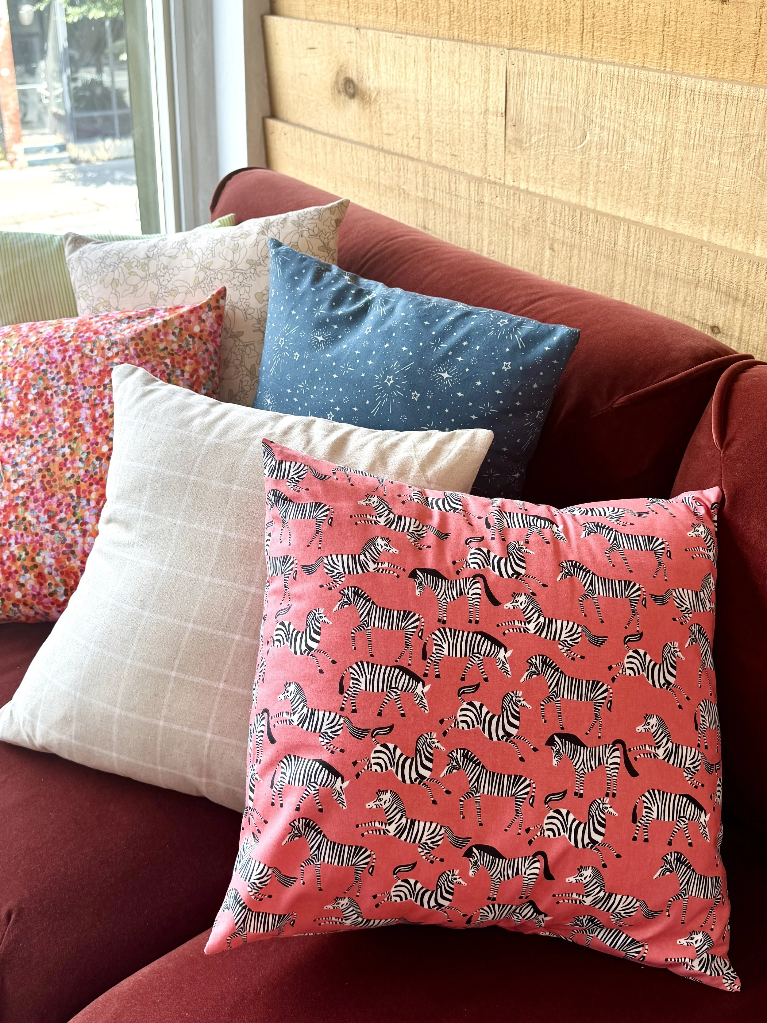 Childrens decorative pillows with colorful patterns and animal prints on red couch in sunlit room.