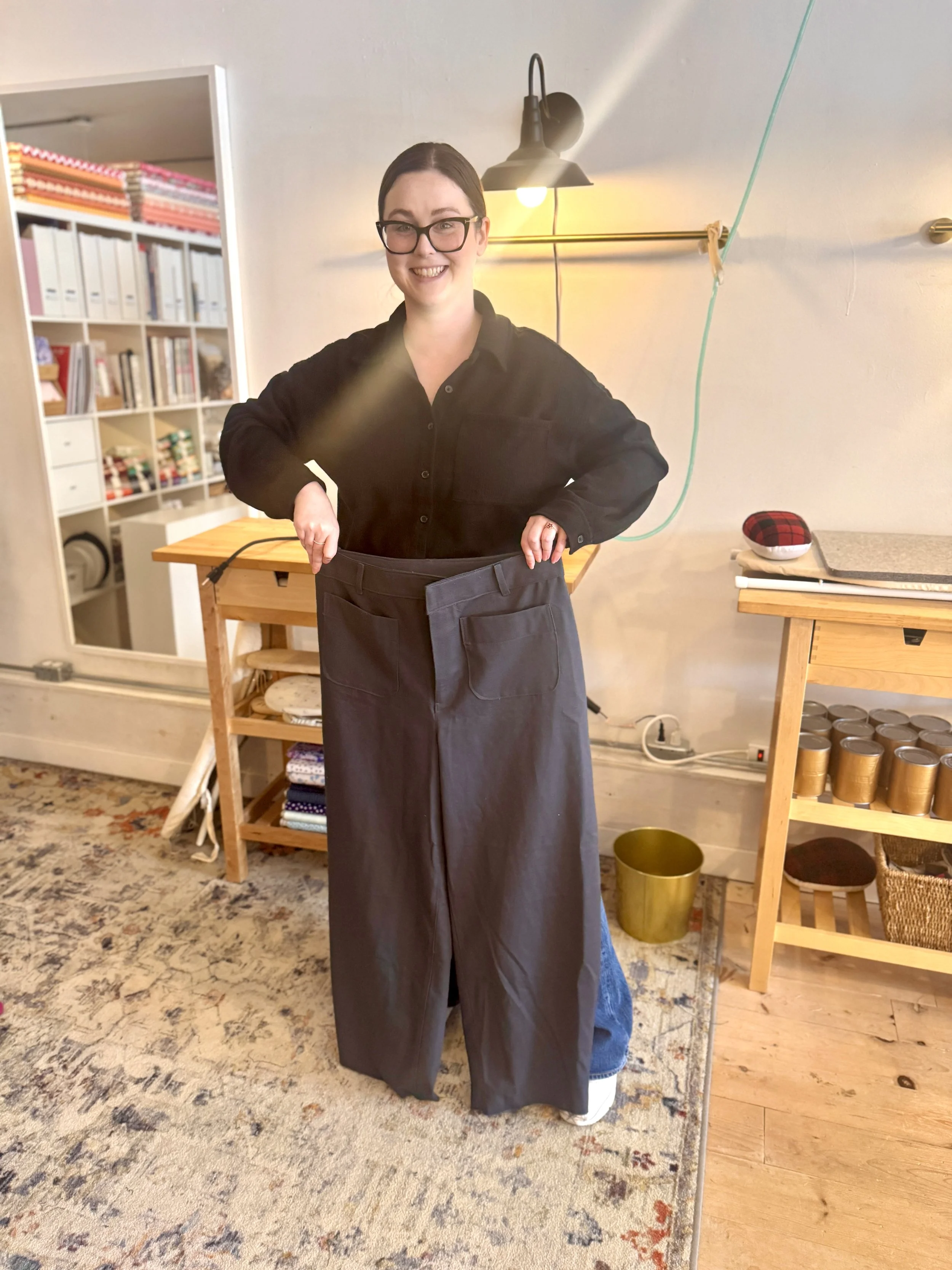 A woman with glasses and a black shirt standing inside, lifting up a pair of oversized black pants while smiling at the camera.