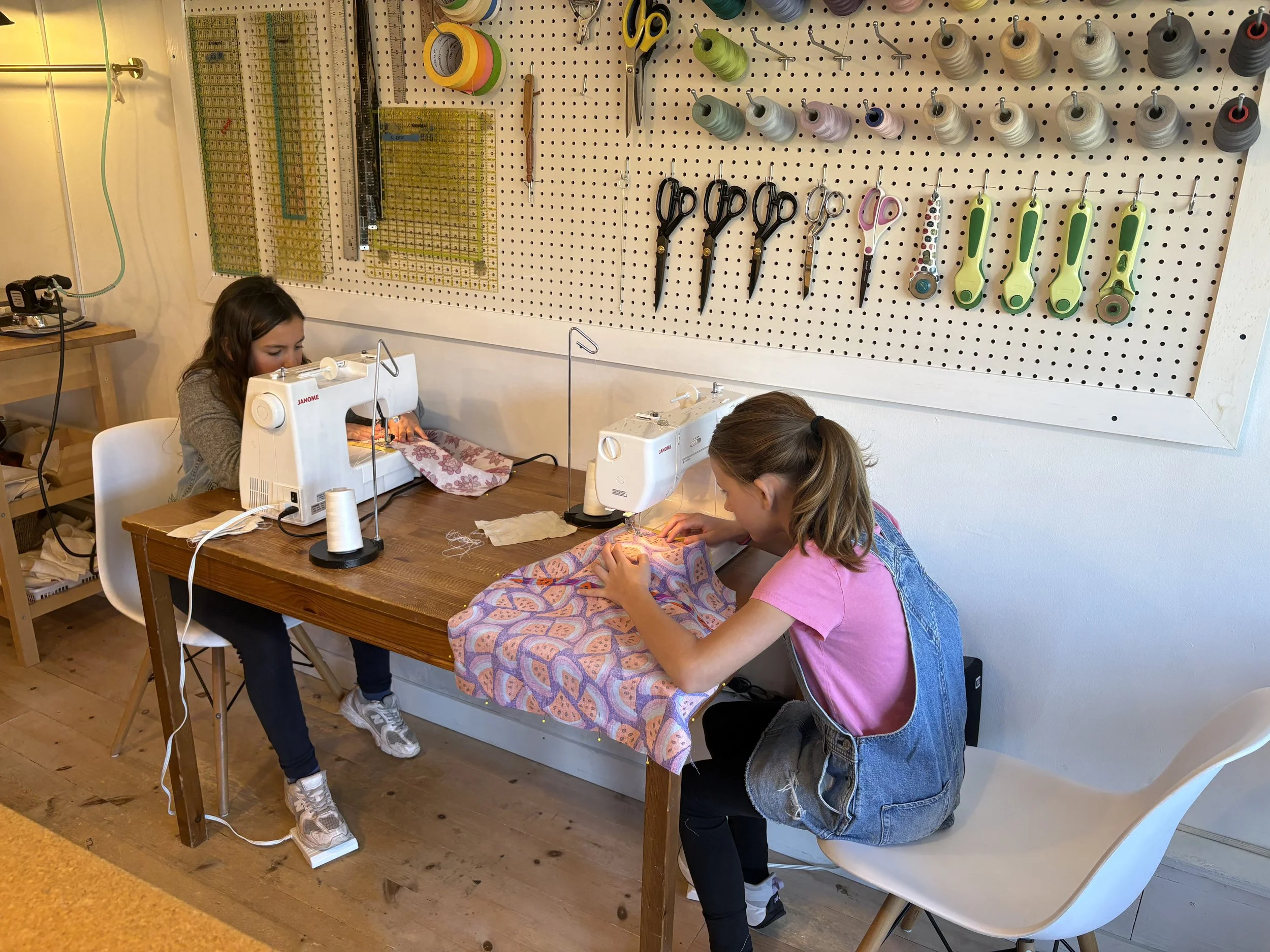 Two young girls working on sewing machines at a workshop table, with sewing tools and fabric, in a craft room with a pegboard full of scissors, scissors, and thread spools on the wall.