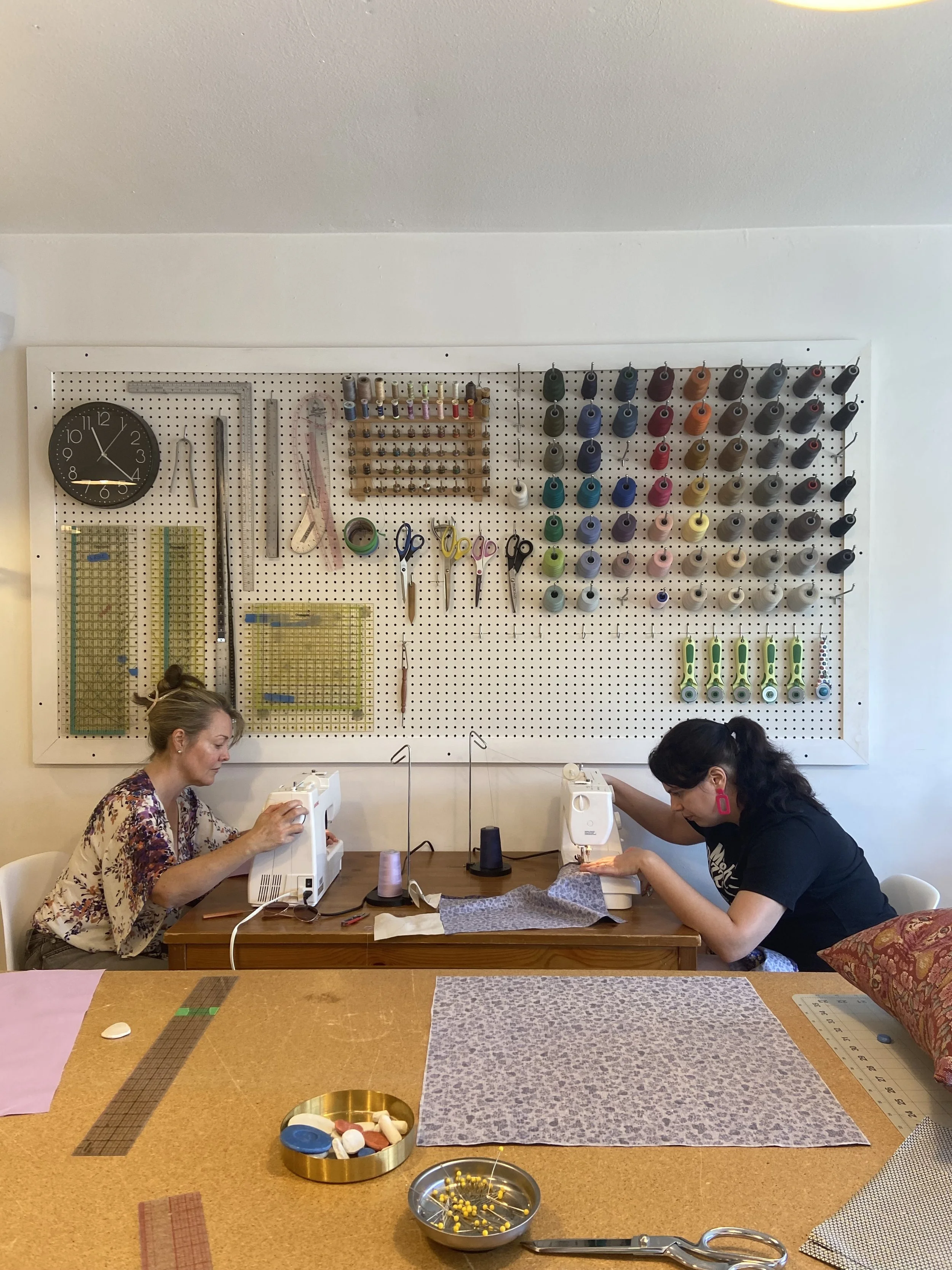 Two women sewing at a table in a craft room with a wall of sewing supplies, including thread spools, scissors, and rulers.