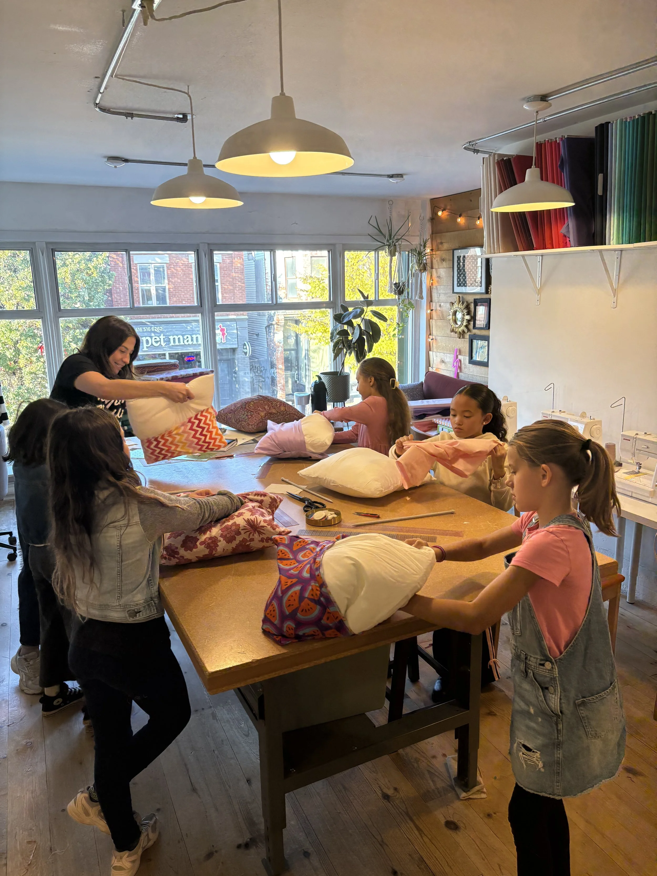 Children and an adult engaging in a pillow fight or craft activity around a large table in a well-lit, cozy room with windows, hanging lights, and shelves of fabric or materials.