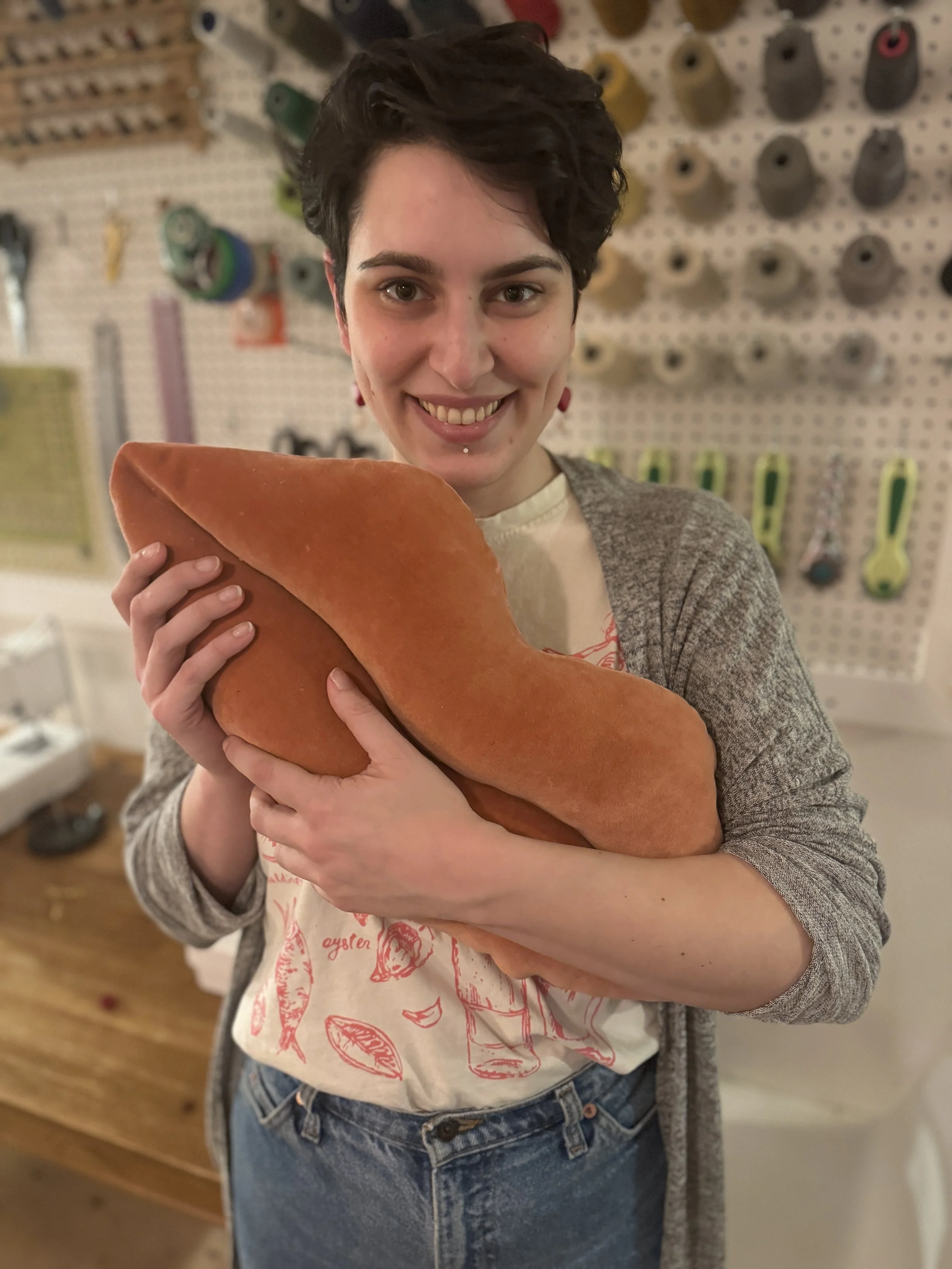 A woman smiling and holding a large loaf of bread in a room with a pegboard wall and various tools in the background.