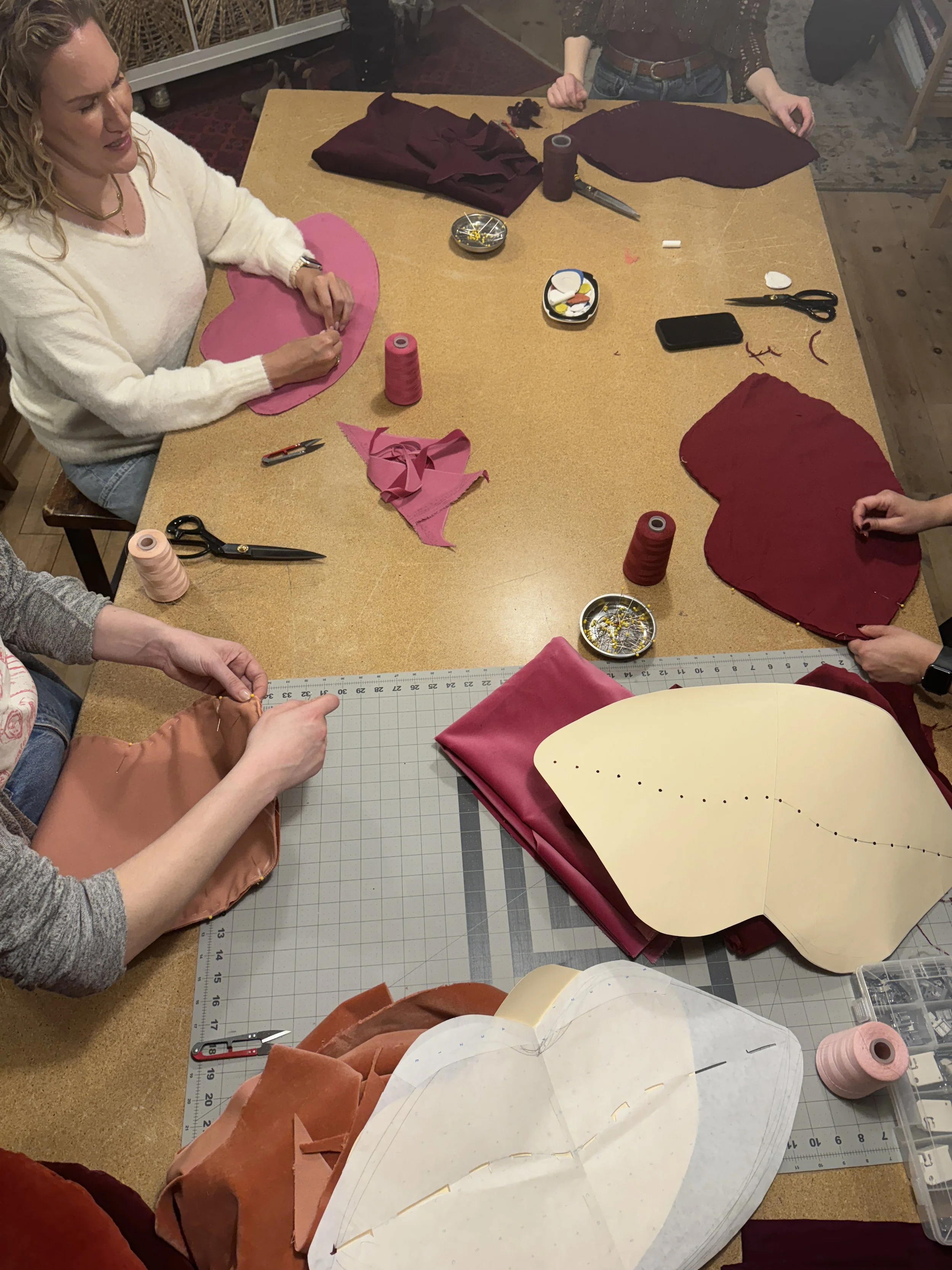 People sitting around a table sewing and assembling fabric, with various fabric pieces, scissors, thread, and sewing supplies.