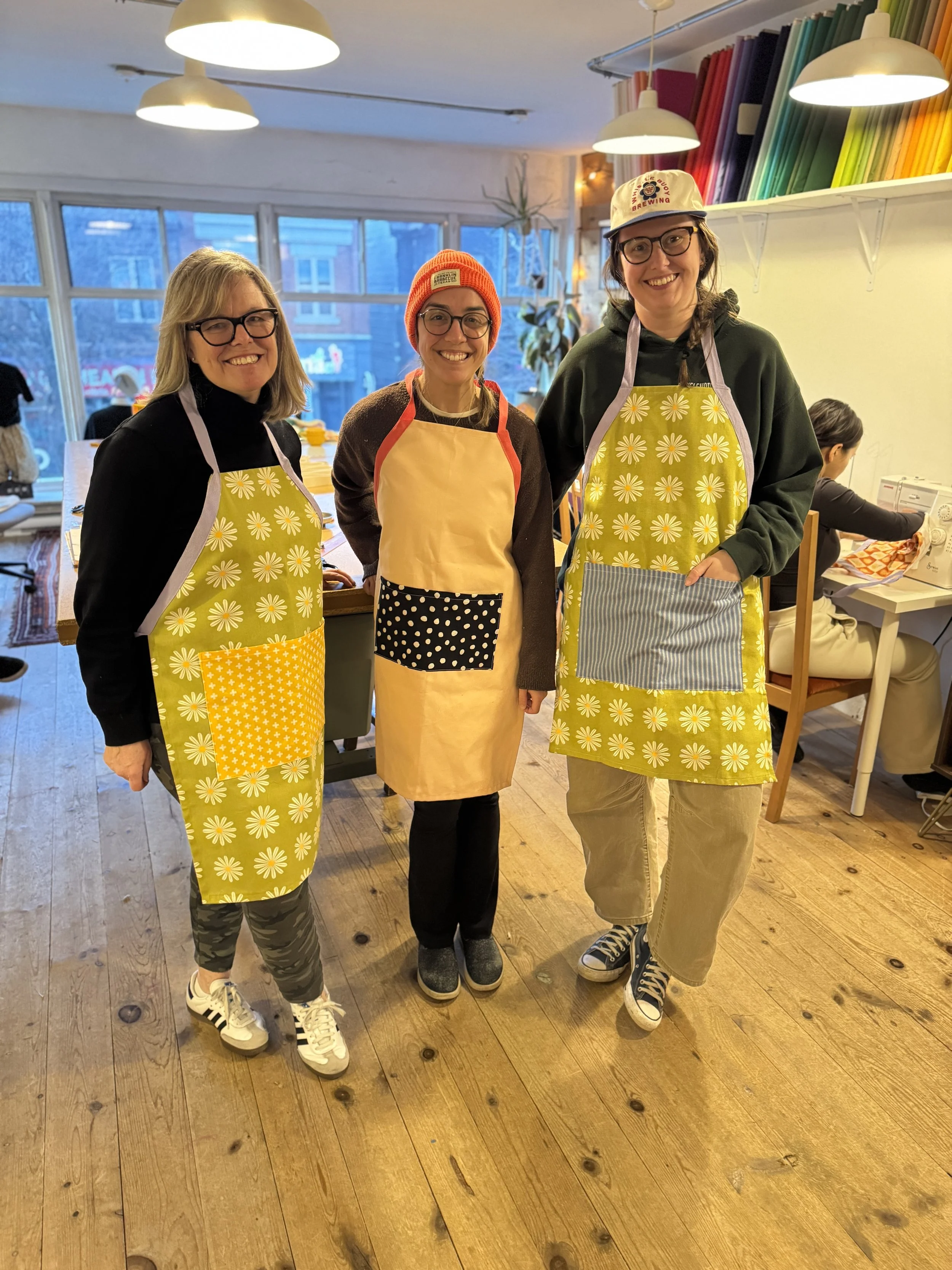 3 students wearing aprons they just made in Learn to Sew 2. Her pet chihuahua sits in the pocket. The apron is full length, made of printed canvas, the print is black 70s style daisies on a beige background. The fabric is a linen and cotton blend. 