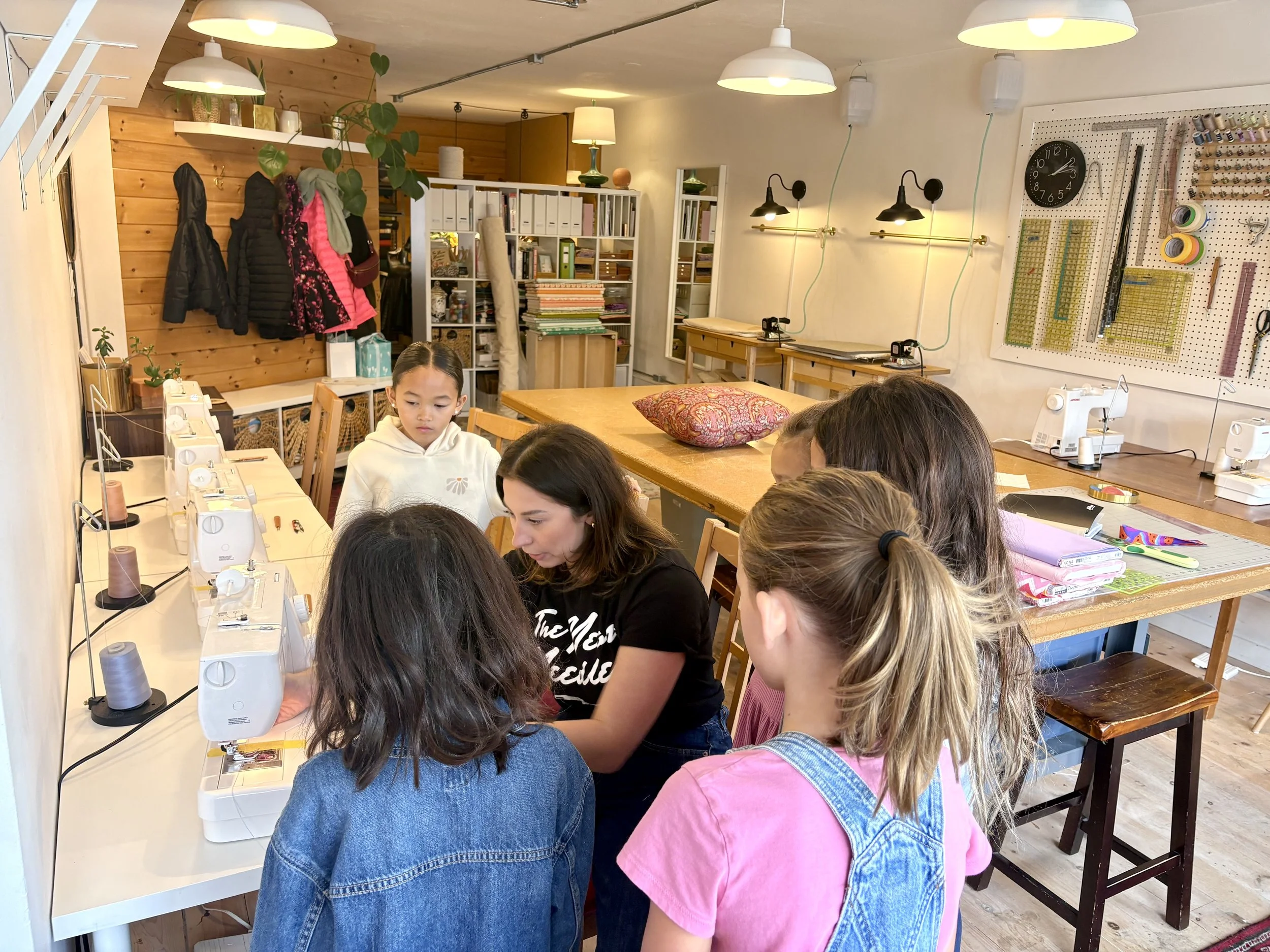 A group of children and an adult woman gathered around multiple sewing machines in a brightly lit crafting room, with shelves holding fabrics, books, and sewing tools in the background.