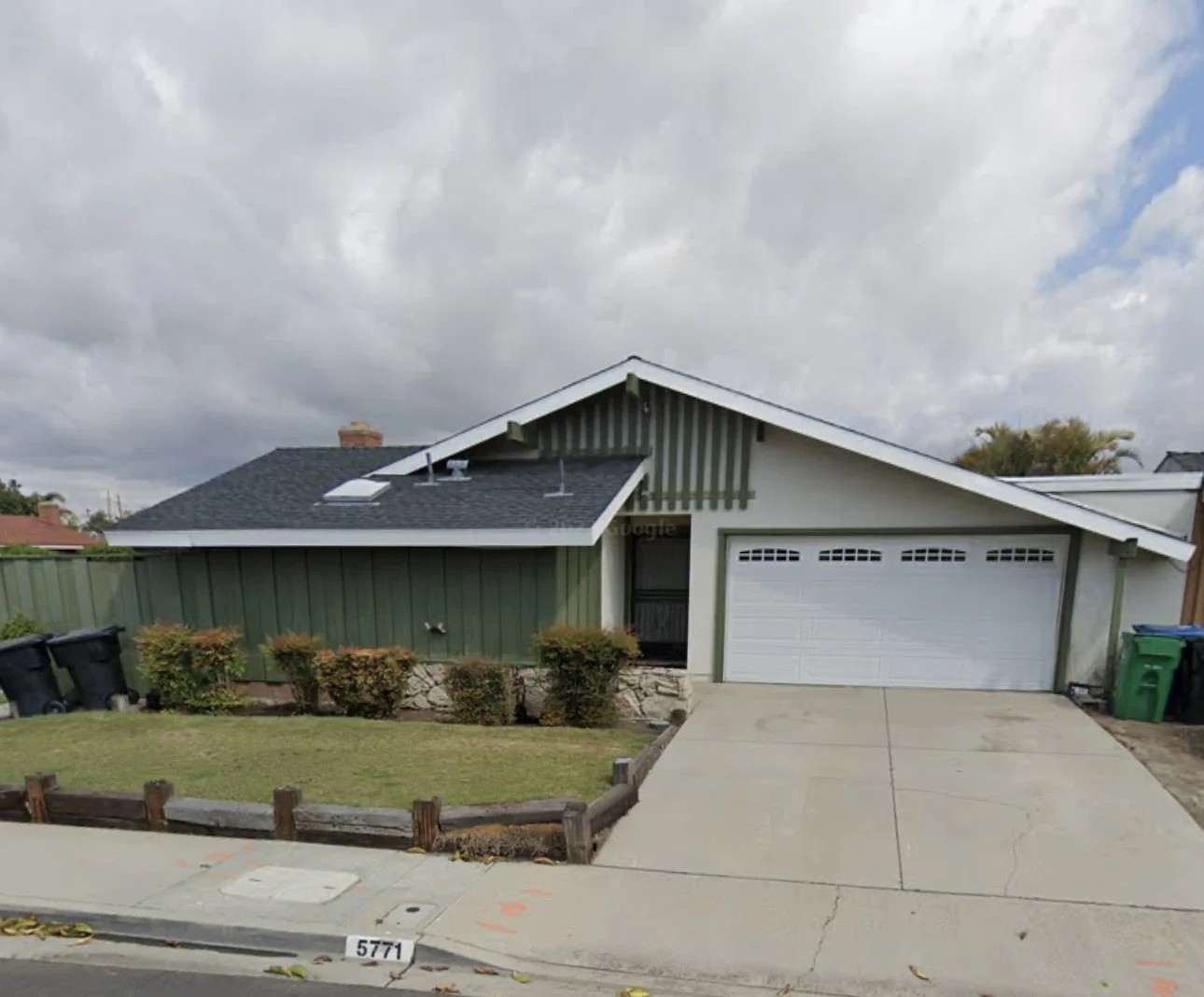 A suburban house with green siding, white garage door, and a sloped roof. There is a small front yard with shrubs and a sidewalk with the house number 5771 visible. The sky is cloudy.