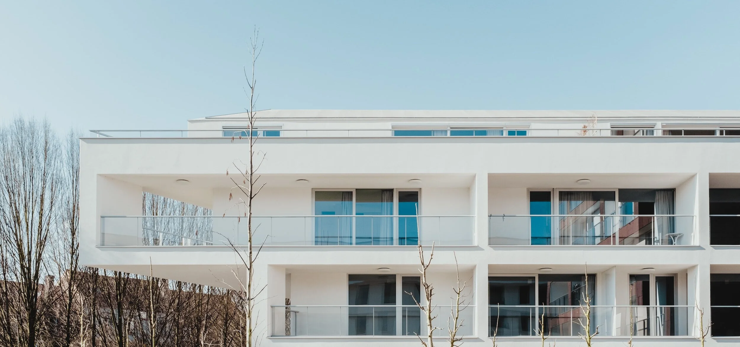 Modern white building with balconies and large windows, trees in the foreground, clear sky.