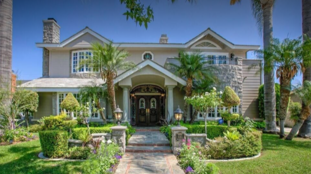 Front view of a large, landscaped house with a stone and siding exterior, arched entrance, and surrounded by palm trees and colorful plants.