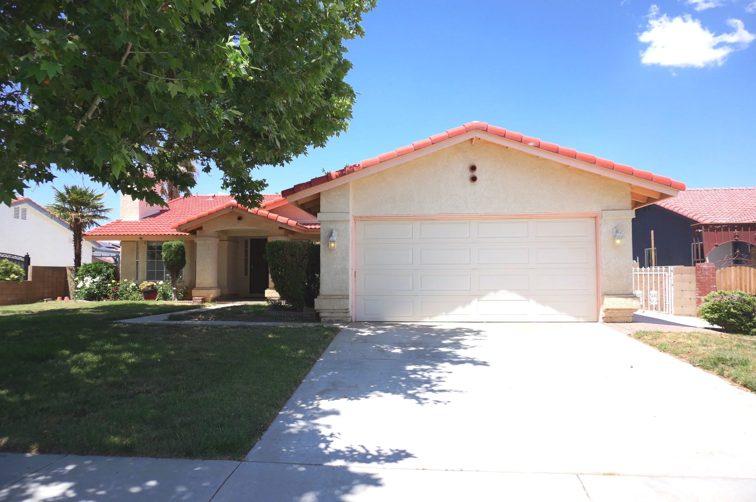 A house with a white garage door, beige exterior walls, and a red-tiled roof. There is a large green tree on the left and a clear blue sky with a few clouds above.