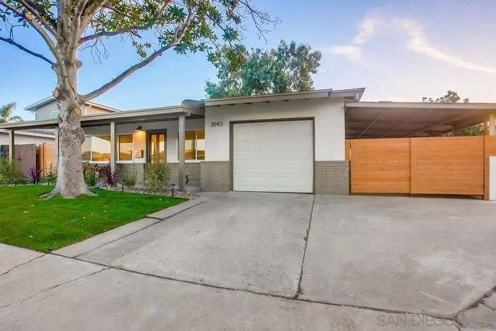 Front view of a mid-century modern house with a concrete driveway, a large tree on the left, and a wooden fence on the right side. The house has a flat roof and a white exterior with large windows.