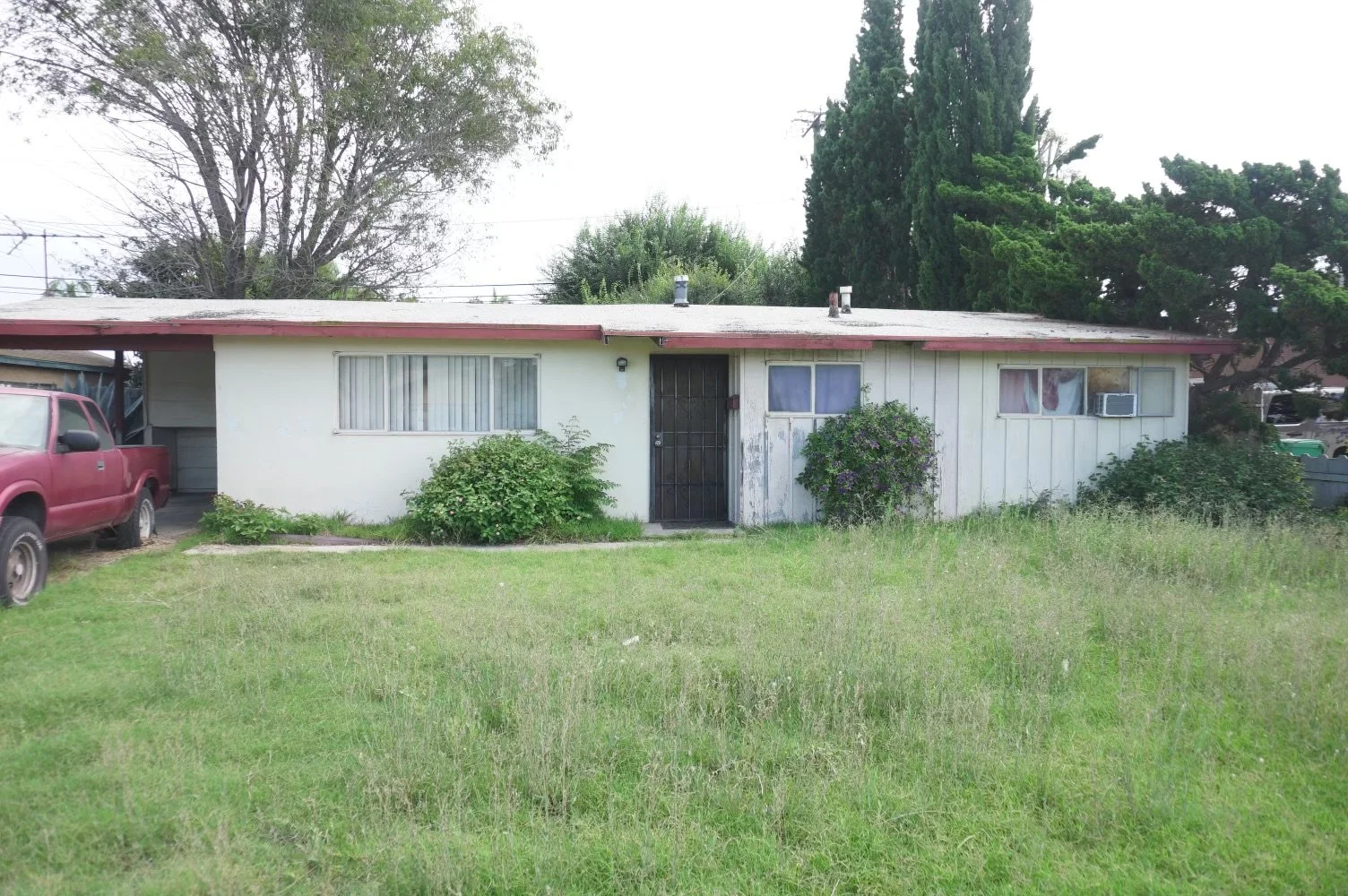 A single-story house with white exterior walls, a red roof, and a front yard with grass, bushes, and some tall trees in the background. There is a red pickup truck parked on the left side.